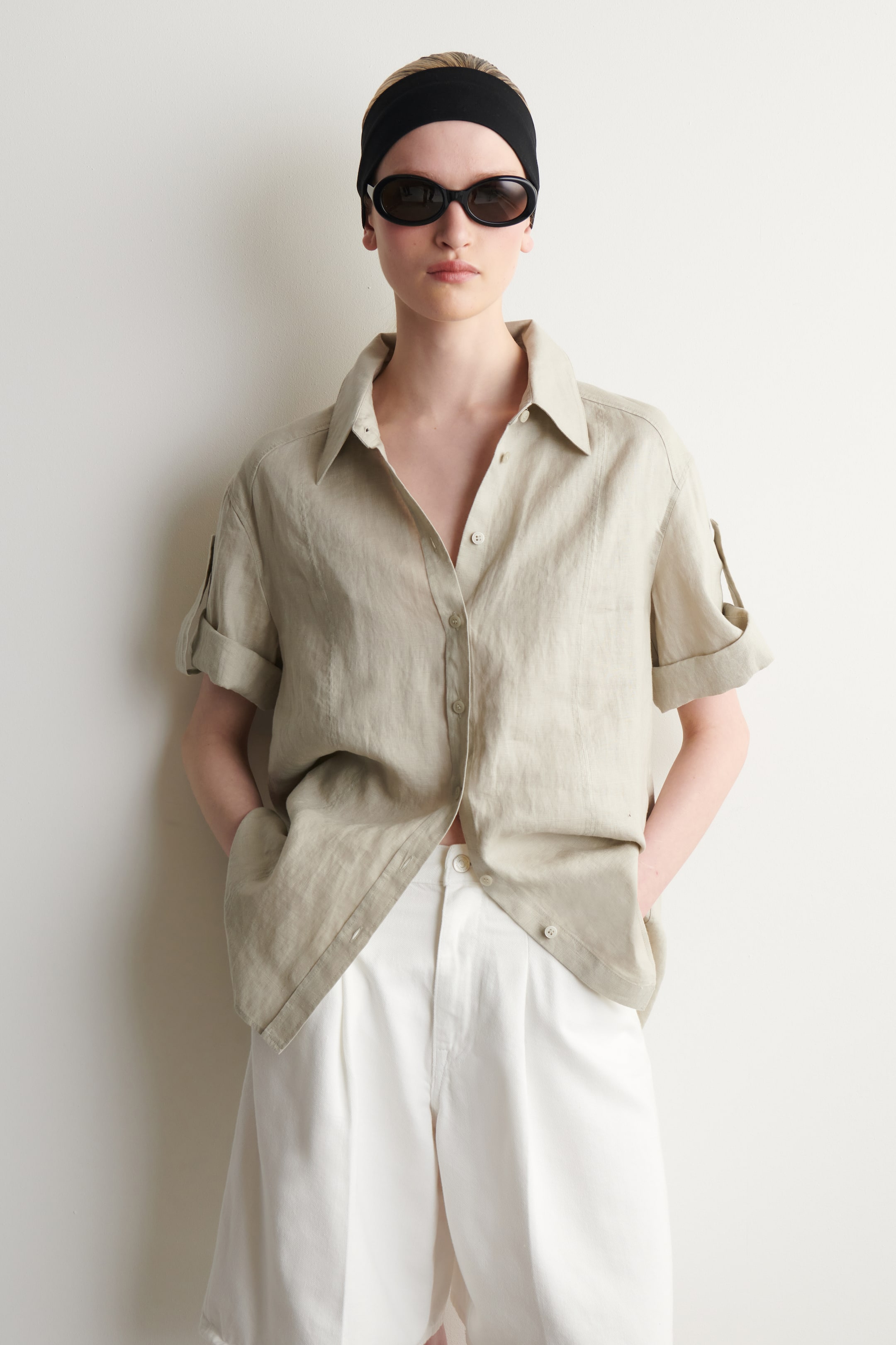 Agrandir l'image: La chemise à manches courtes en lin beige clair est portée sous un léger angle frontal, mettant en valeur sa coupe décontractée et son tombé. Elle présente un col évasé, des poignets retroussés, et est partiellement déboutonnée tout en étant rentrée dans un short blanc plissé.