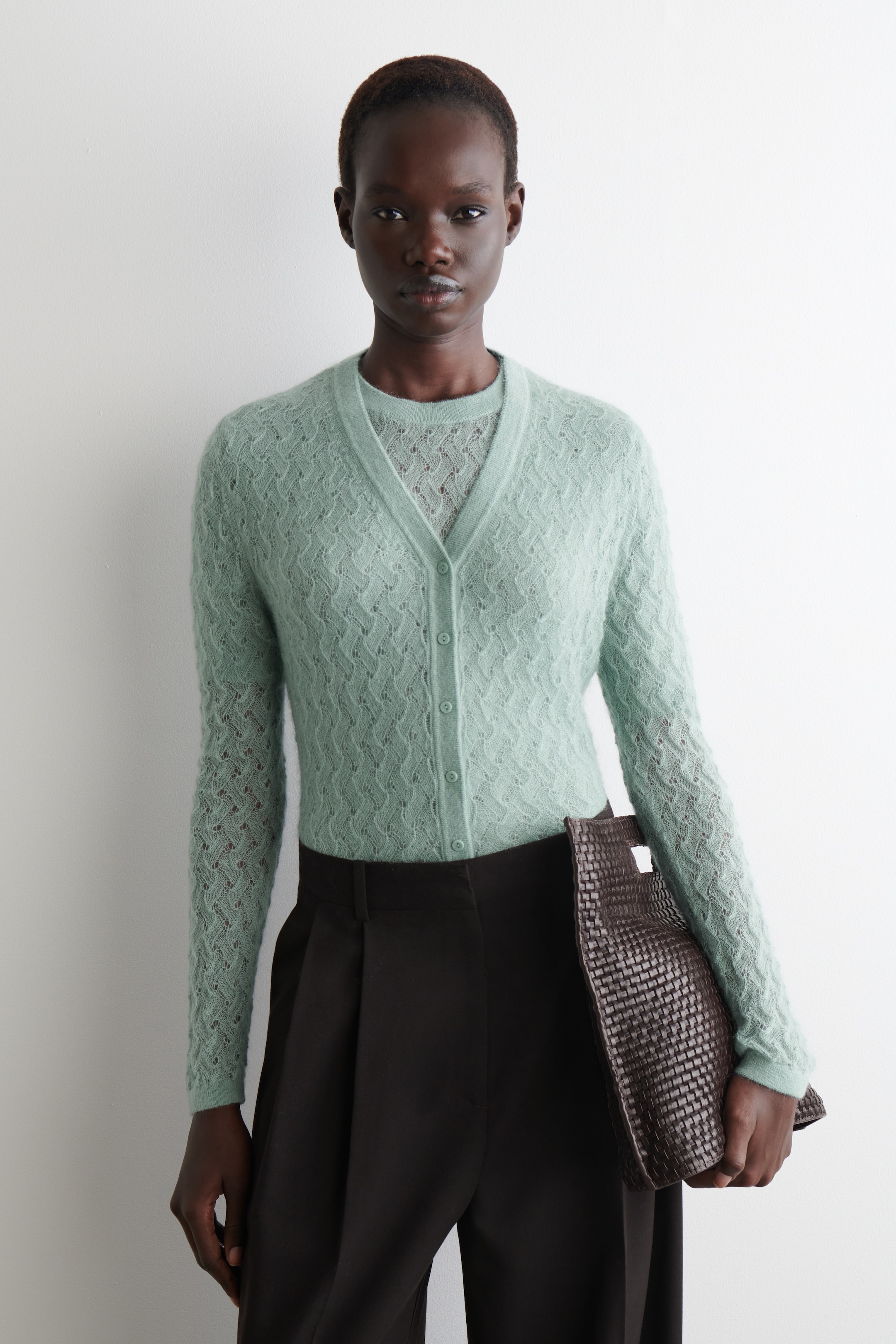 Grösseres Bild ansehen: Eine hellgrüne V-Ausschnitt-Strickjacke mit einem zarten offenen Strickmuster und einer Knopfleiste wird über einem passenden Rundhalstop getragen. Die langen Ärmel der Strickjacke und die flauschige Textur sind sichtbar. Dunkle, plissierte, weit geschnittene Hosen werden getragen, und eine dunkelbraune geflochtene Tasche wird gehalten.