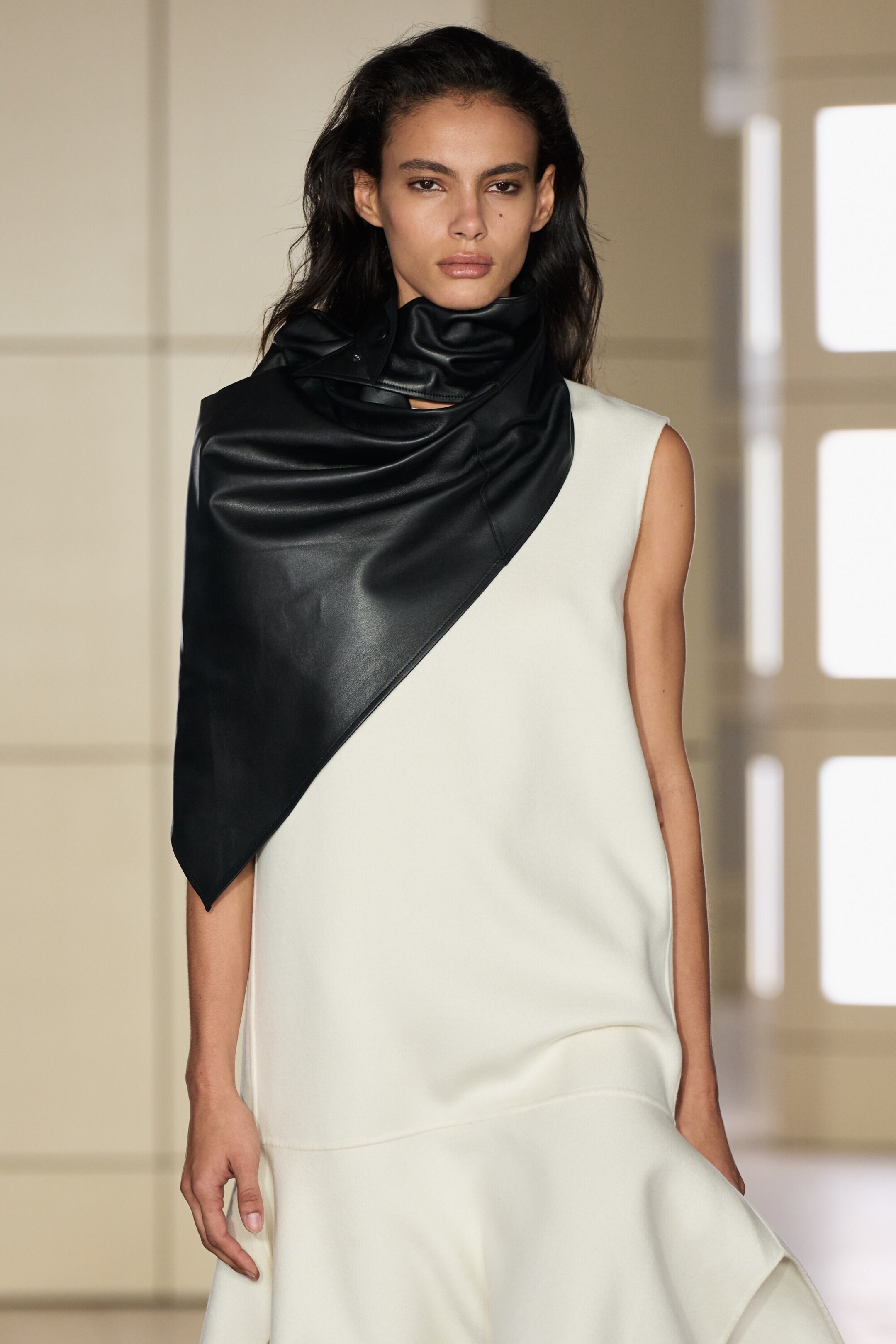 A black leather hood drapes asymmetrically over a sleeveless cream dress, facing front to showcase its structured design. The accessory wraps snugly around the neck in a gathered cowl style and extends diagonally across the chest and one shoulder in a smooth, triangular shape. A black leather hood drapes asymmetrically over a sleeveless cream dress, facing front to showcase its structured design. The accessory wraps snugly around the neck in a gathered cowl style and extends diagonally across the chest and one shoulder in a smooth, triangular shape.