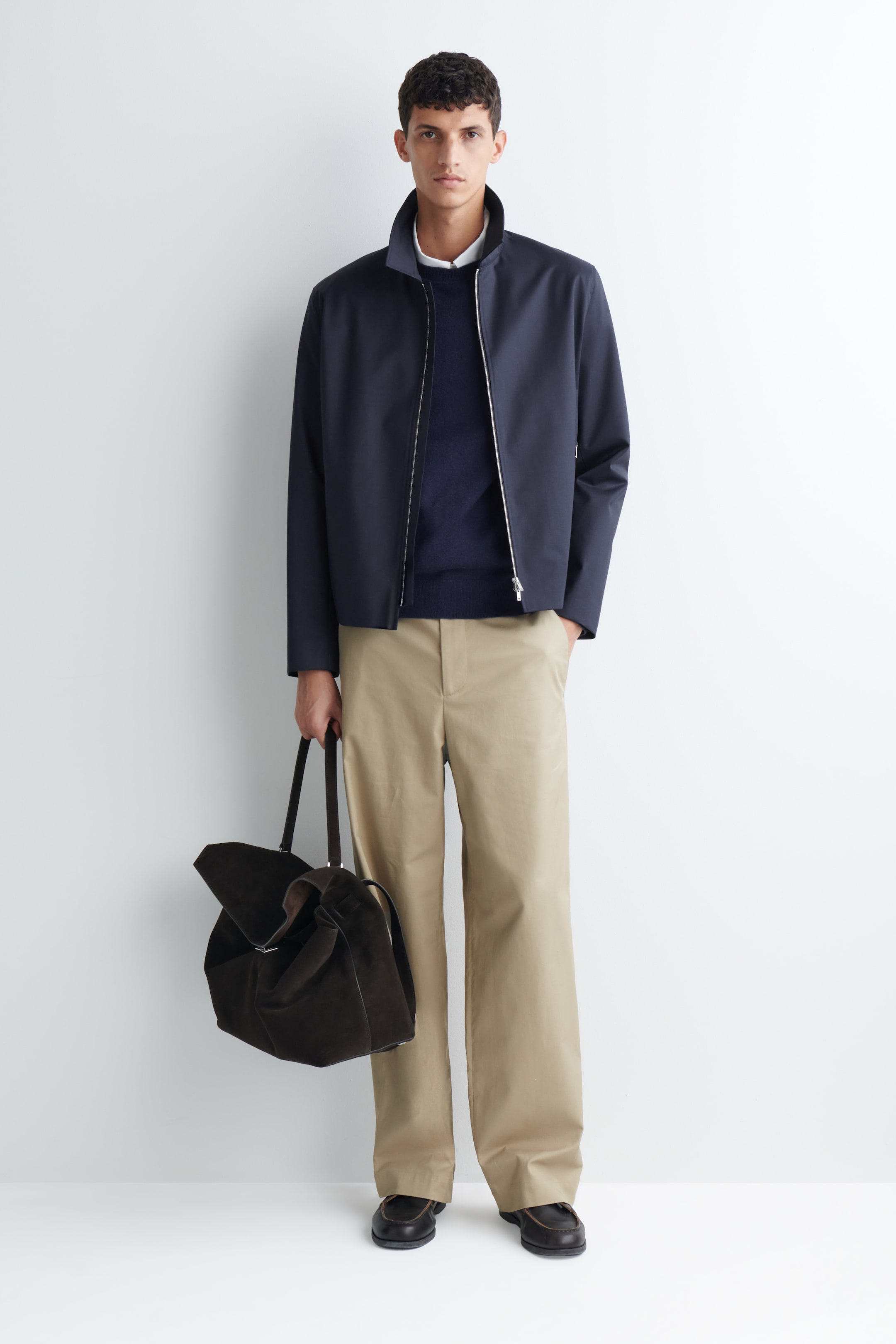 Visa större bild: Dessa beige chinos i bomull, med en avslappnad vid benvidd och en ren linning, bärs av modellen. De är stylade med en marinblå jacka med dragkedja, en mörk stickad tröja och mörka loafers, medan modellen håller i en mörkbrun väska i mocka.