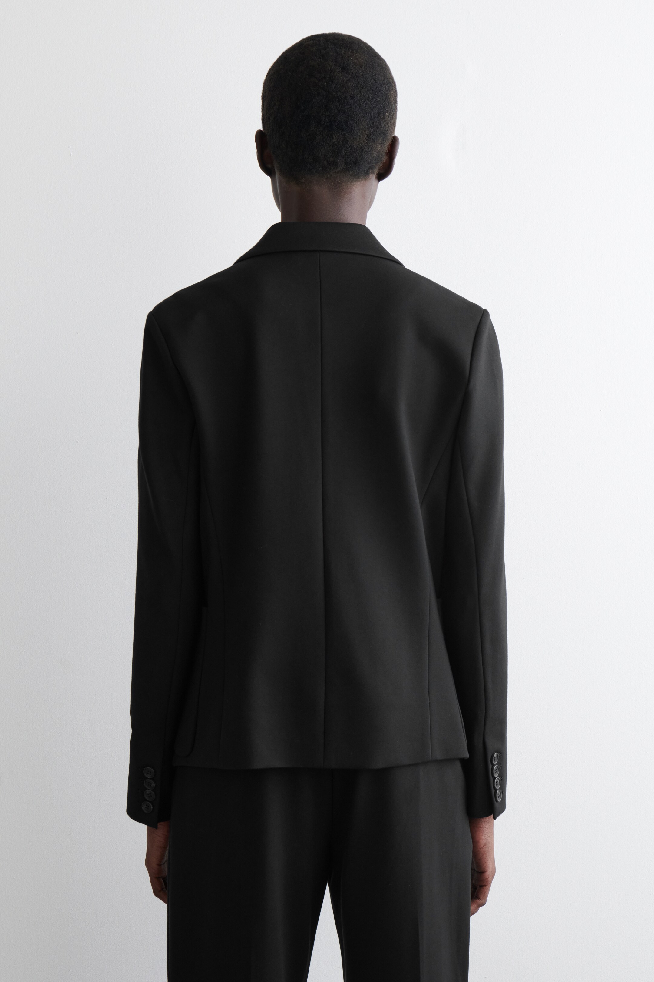 Zobrazit větší obrázek: Černé sako je prezentováno zezadu, ukazující jeho strukturovanou siluetu, jediný středový zadní šev a čtyři knoflíky na každé manžetě rukávu. Odpovídající černé kalhoty jsou také nošeny, dokončující monochromatický celek.