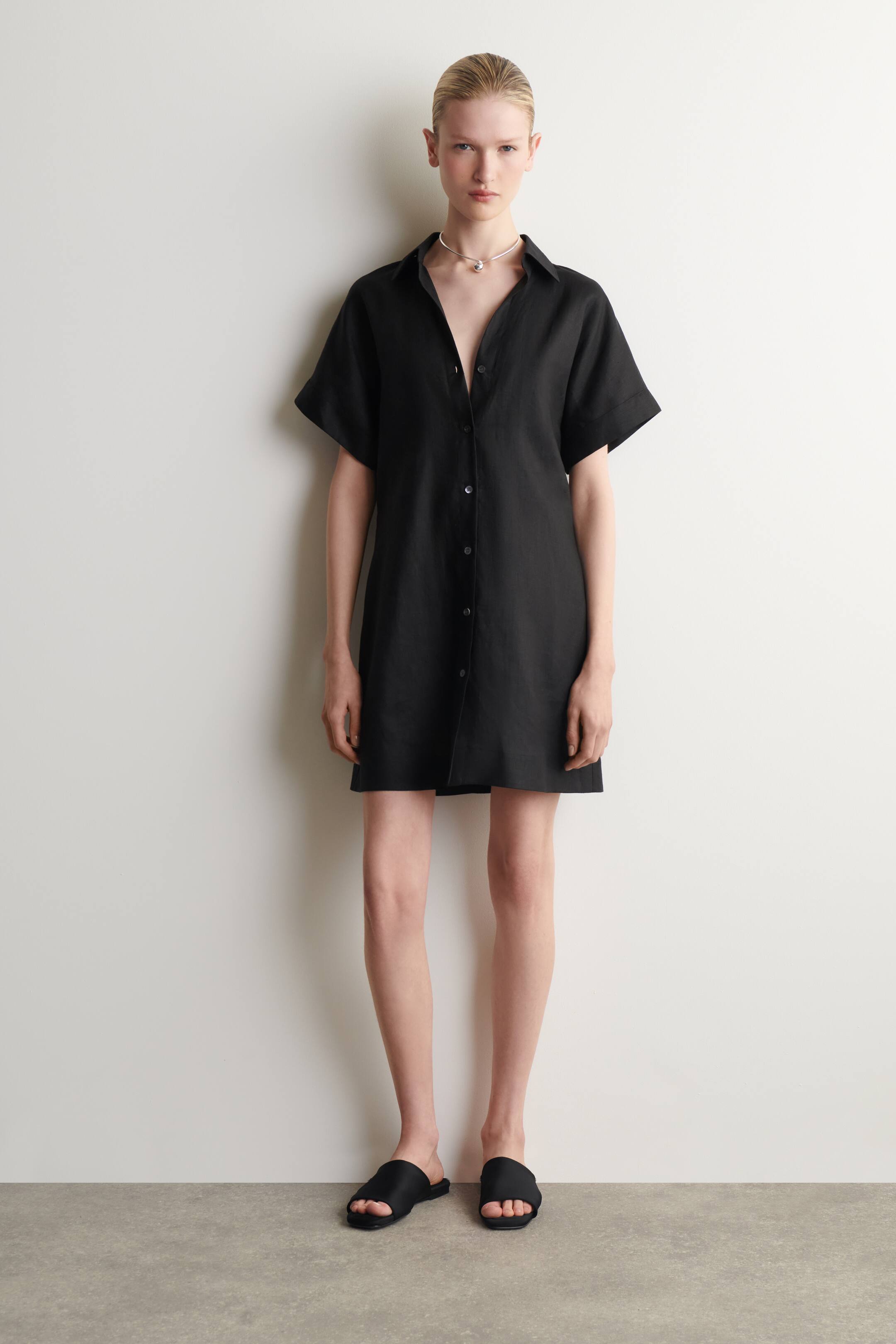 Ver imagen más grande: Un vestido camisero negro con textura presenta un ajuste relajado y de longitud mini. Cuenta con un cuello en punta, mangas cortas y anchas con puños doblados, y una botonadura completa en el frente con algunos botones desabrochados en la parte superior.