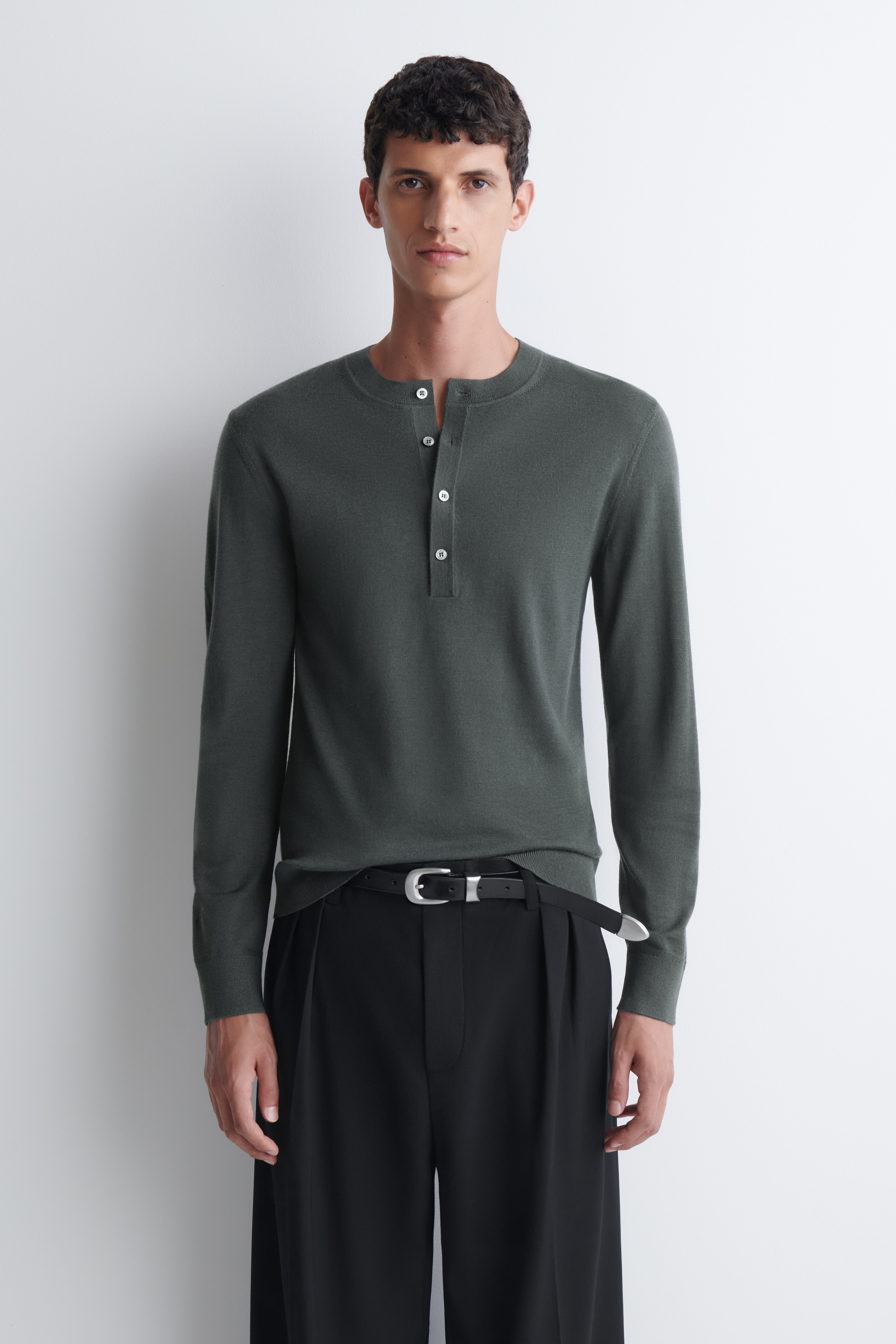 Un top henley slim in lana merino verde scuro Ã¨ mostrato frontalmente, con girocollo a coste, abbottonatura a quattro bottoni e maniche lunghe. Il top Ã¨ rifinito con polsini a coste e orlo a coste.