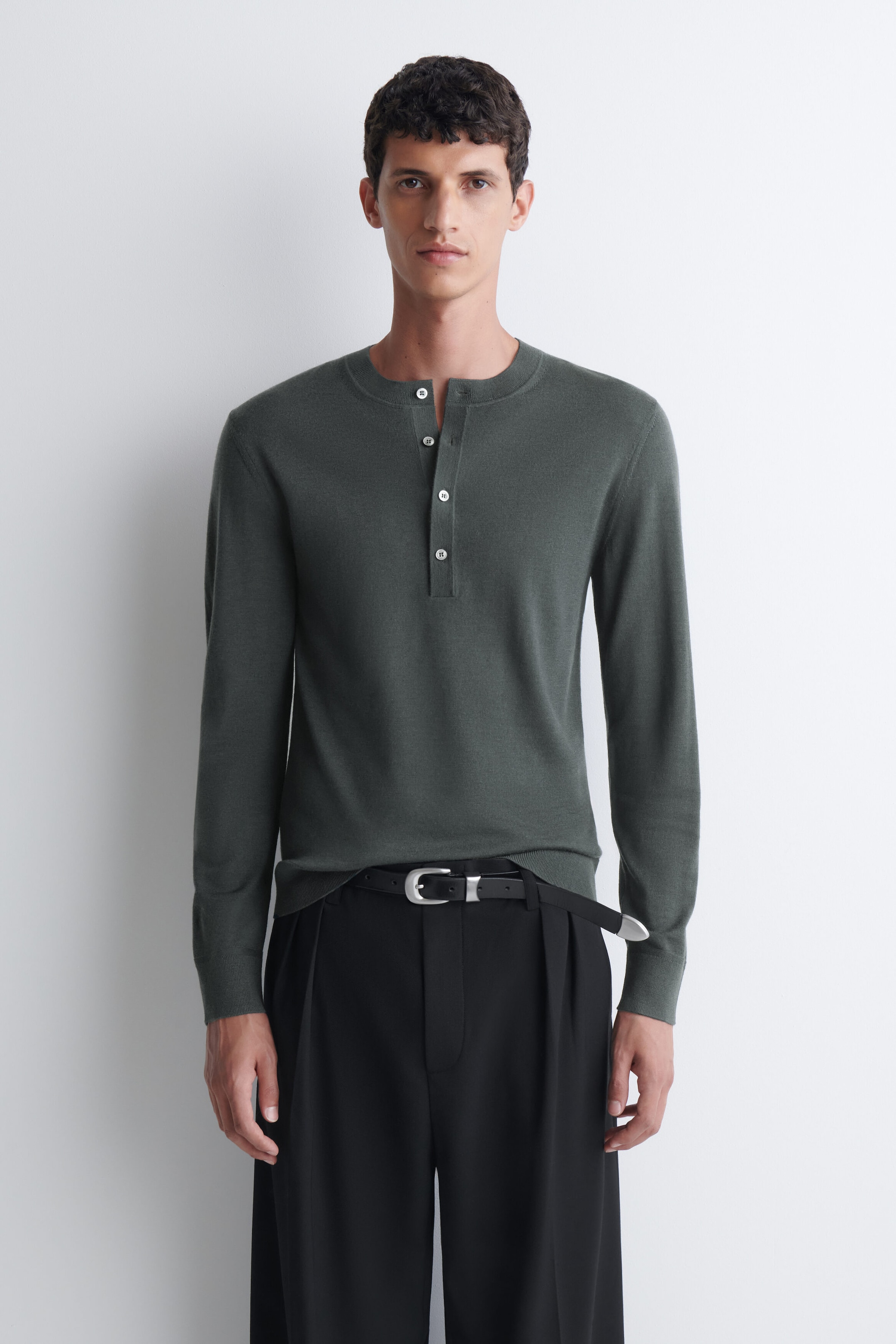 A slim-fit, dark green merino wool henley top with long sleeves and a partially unbuttoned placket featuring contrasting white buttons is worn by the model. The top is styled tucked into black pleated trousers with a black belt featuring a silver buckle.