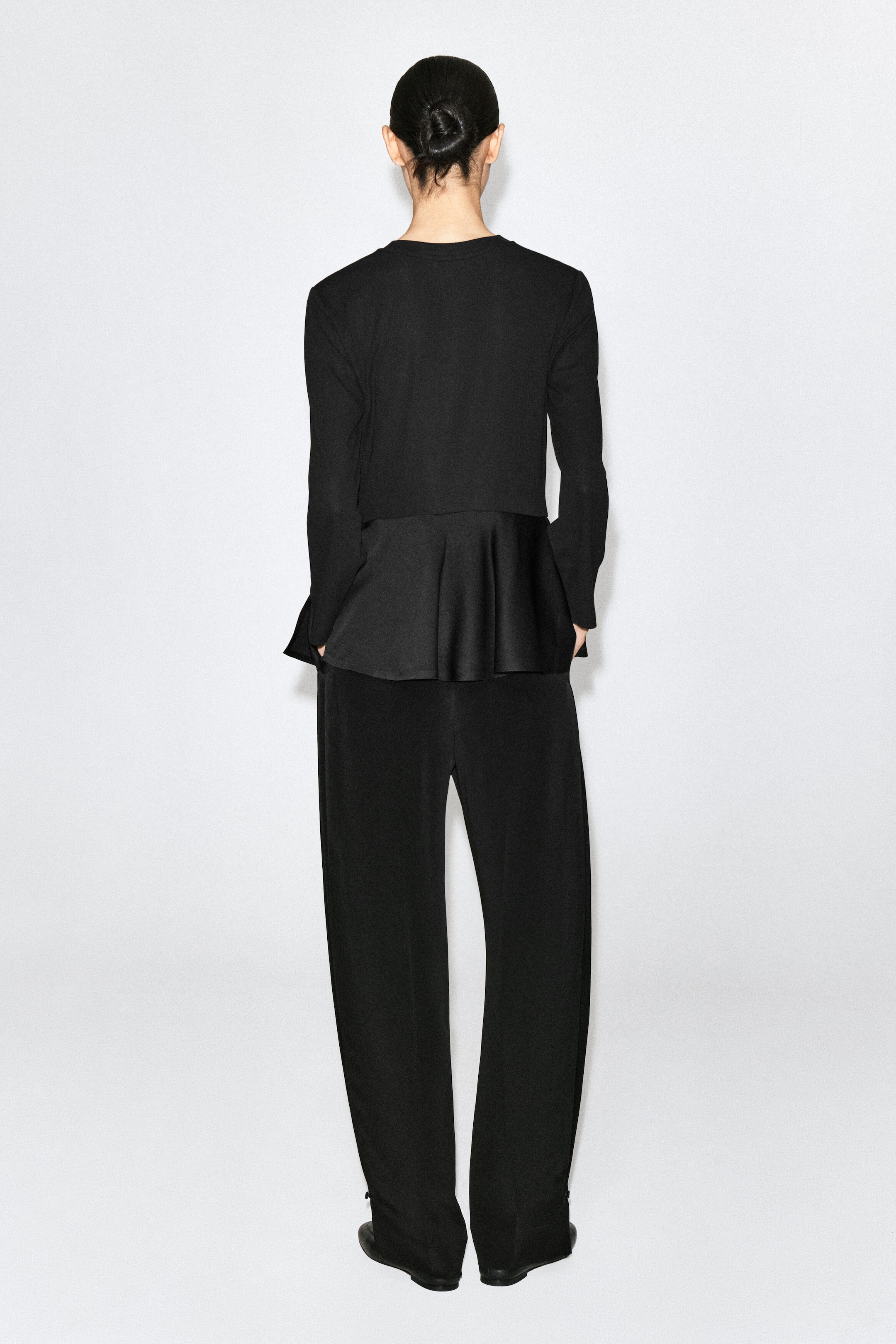 Un haut en jersey noir à basque en satin est porté par le mannequin, vu de dos. Le haut à manches longues présente une partie supérieure en tissu mat et un ourlet basque évasé en satin, agencé avec un pantalon noir assorti.