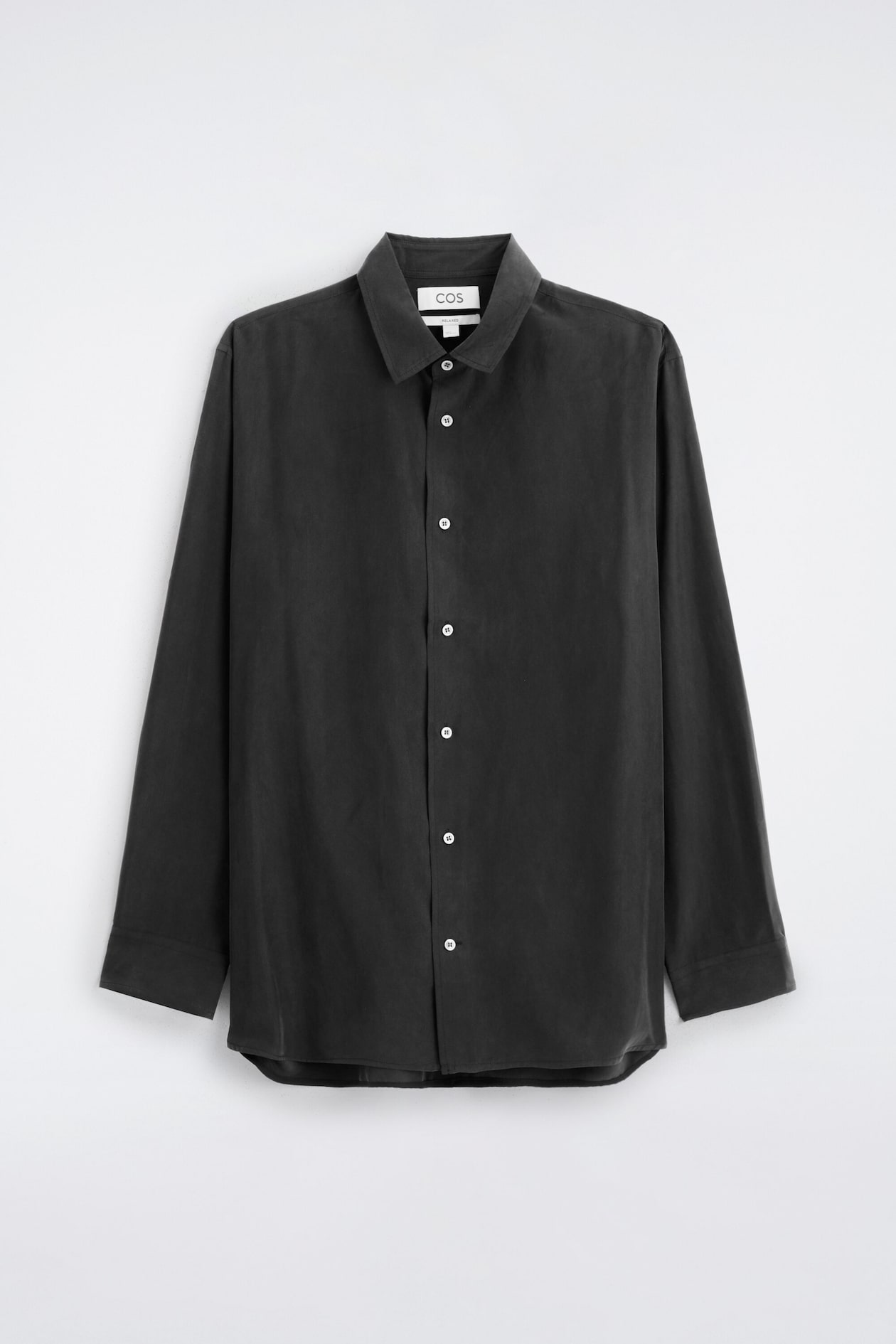 A sand-washed silk shirt in dark grey is shown from the front, featuring a classic point collar, a button-down front with contrasting light-coloured buttons, and long sleeves. The shirt is shown in a relaxed fit with a soft, matte texture and a slightly curved hem.