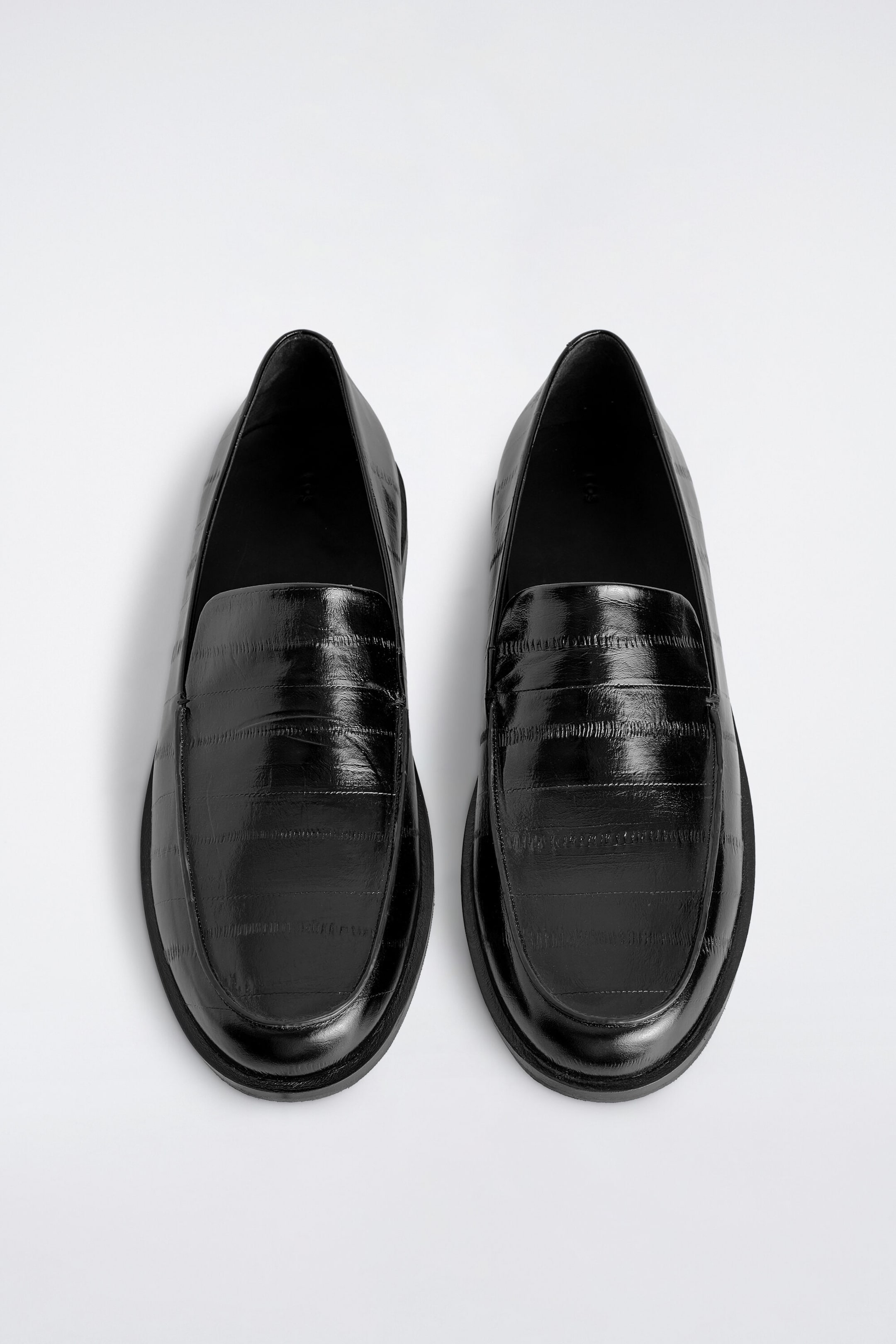 A pair of black polished leather loafers is laid flat in a top-down view, showcasing a distinctive eel-effect embossed texture. The loafers feature rounded toes, a saddle strap across the vamp, and low-profile black soles. A pair of black polished leather loafers is laid flat in a top-down view, showcasing a distinctive eel-effect embossed texture. The loafers feature rounded toes, a saddle strap across the vamp, and low-profile black soles.