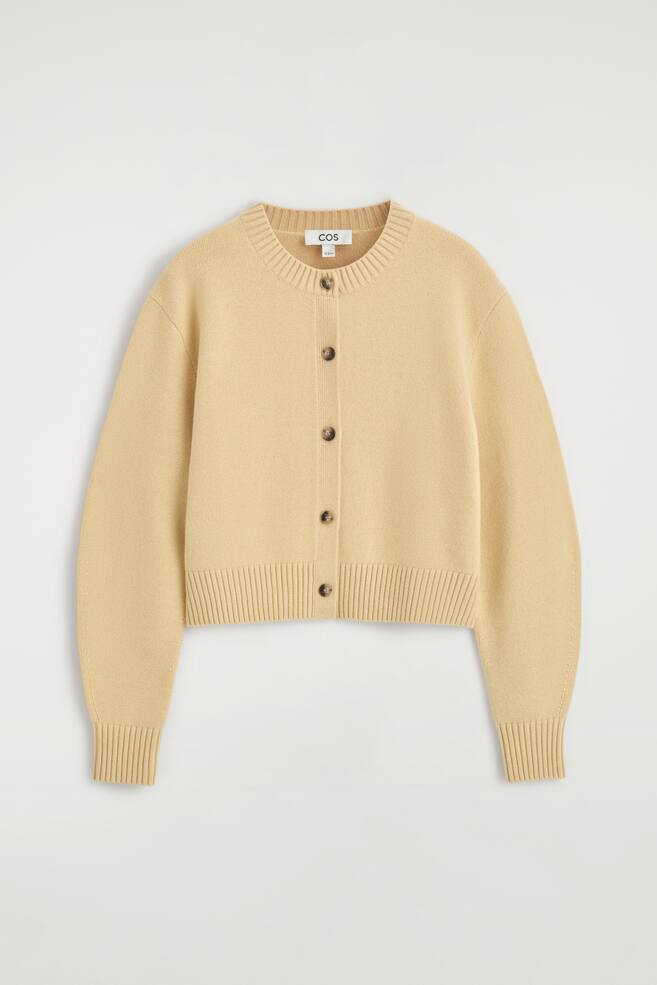 En lysegul cardigan i merinould set forfra, med rund hals, knaplukning foran med fem mÃ¸rke knapper i skildpaddeskalslook og sÃ¦nkede skuldre. Den kortklippede cardigan fremviser en bred ribkant forneden og matchende ribmanchetter. - 1