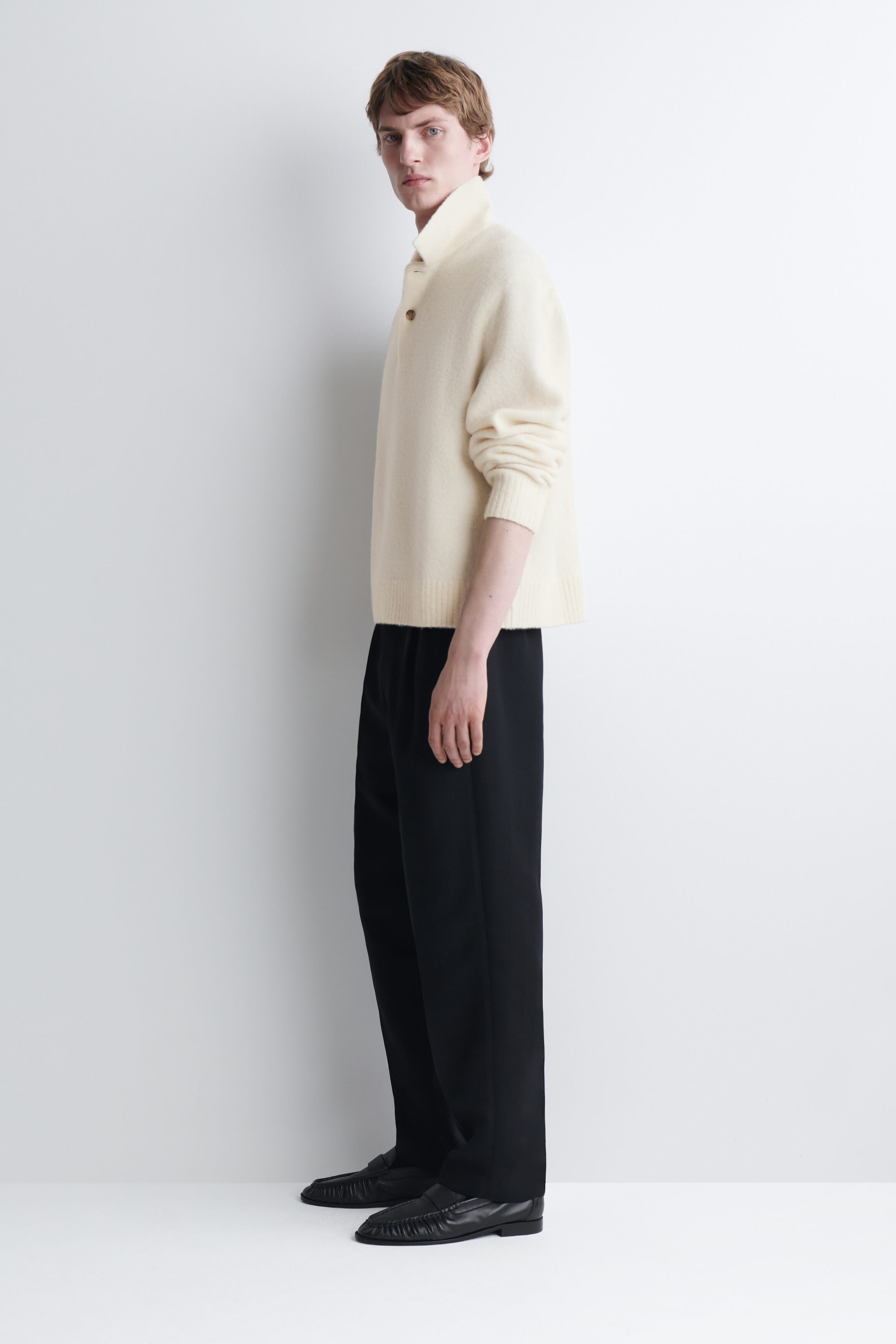 A cream-colored brushed wool polo shirt is worn by the model, featuring a pointed collar, a buttoned placket, and ribbed cuffs and hem. The shirt is styled with black trousers and black loafers.