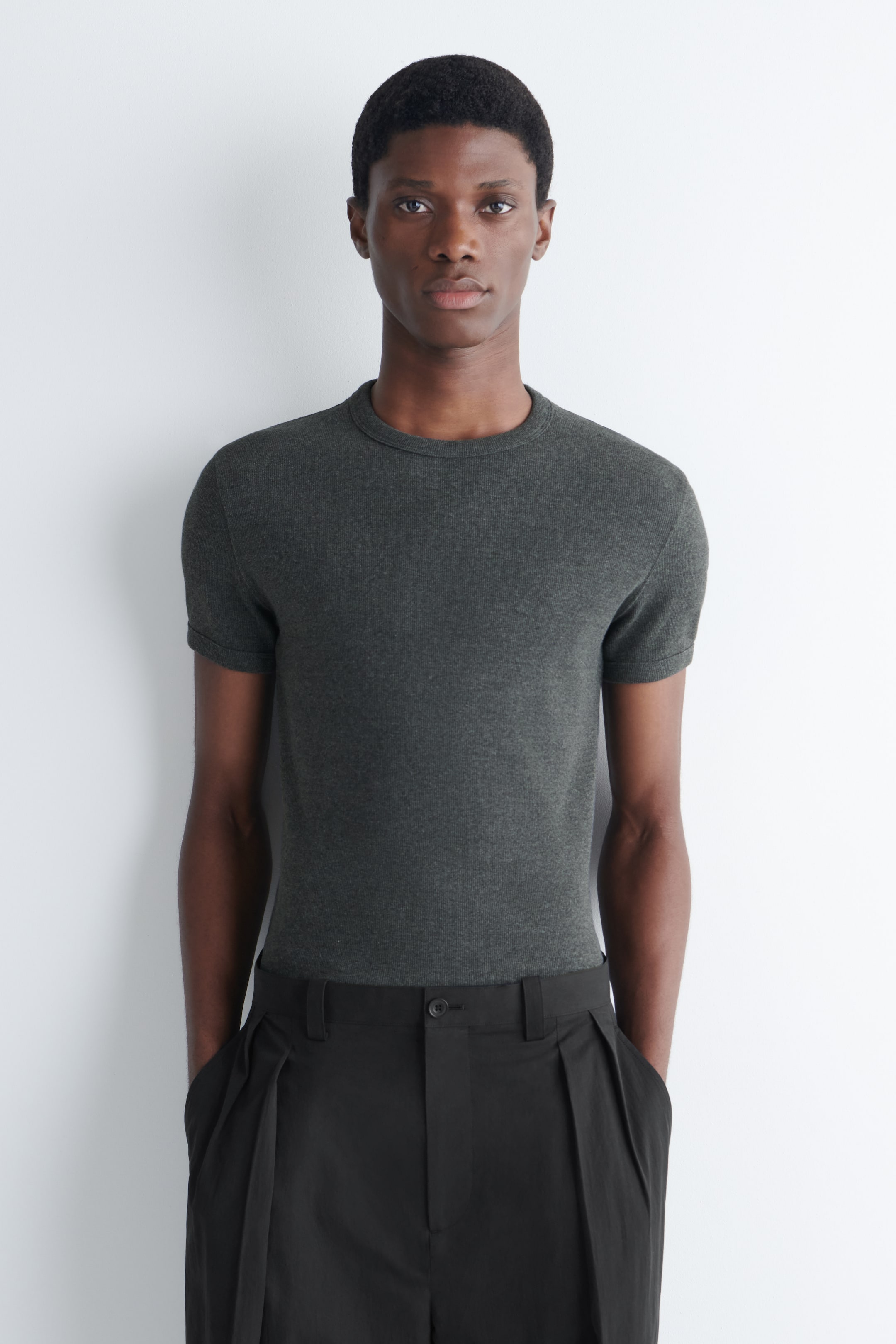 View larger image: A dark grey short-sleeved top featuring a fine ribbed knit texture and a crew neck. It fits closely to the body and is tucked into black trousers.