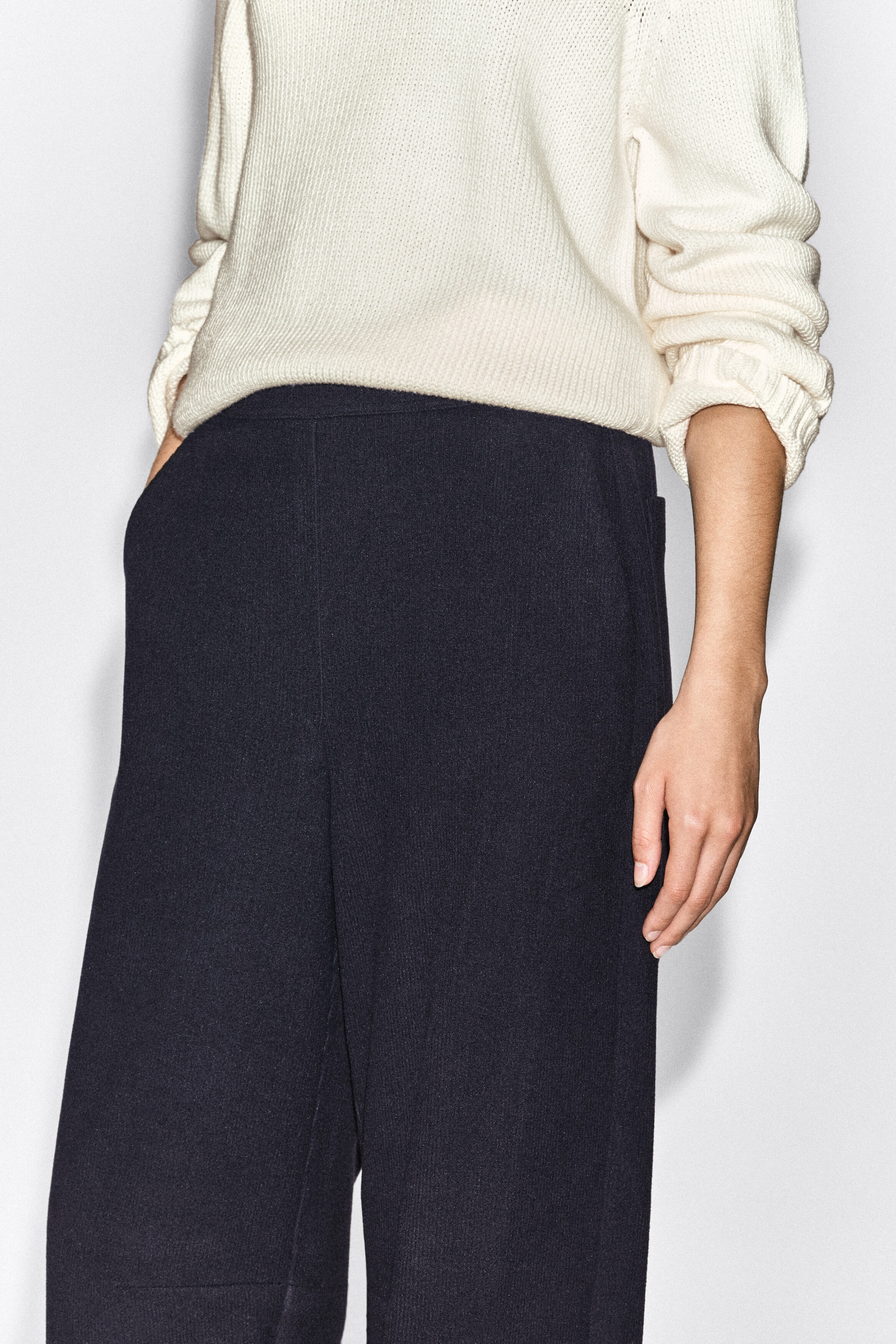 Corduroy-jersey wide-leg pants in dark navy are worn by the model, who has one hand resting in a side pocket. The pants feature a fine-ribbed texture and are styled with a cream knit sweater tucked into the waistband.