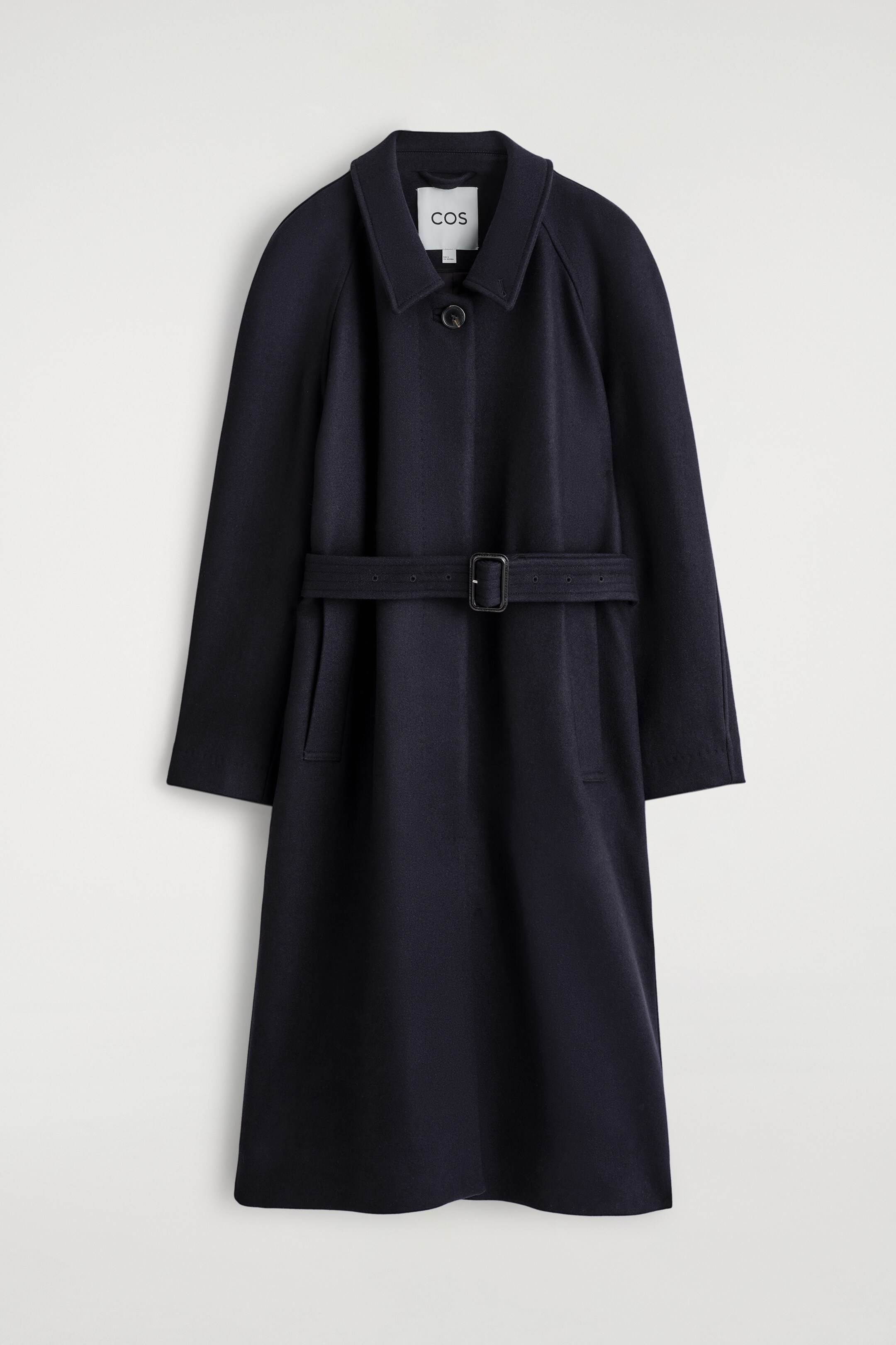 Ingrandisci l'immagine: Un cappotto blu navy scuro è mostrato dal davanti, caratterizzato da una silhouette rilassata e una lunghezza lunga. È progettato con un colletto a punta che mostra un'etichetta del marchio visibile, maniche raglan, una chiusura con un solo bottone al collo, una cintura in tessuto abbinato con una fibbia rettangolare scura che stringe la vita e due tasche a filetto verticali sotto la cintura.
