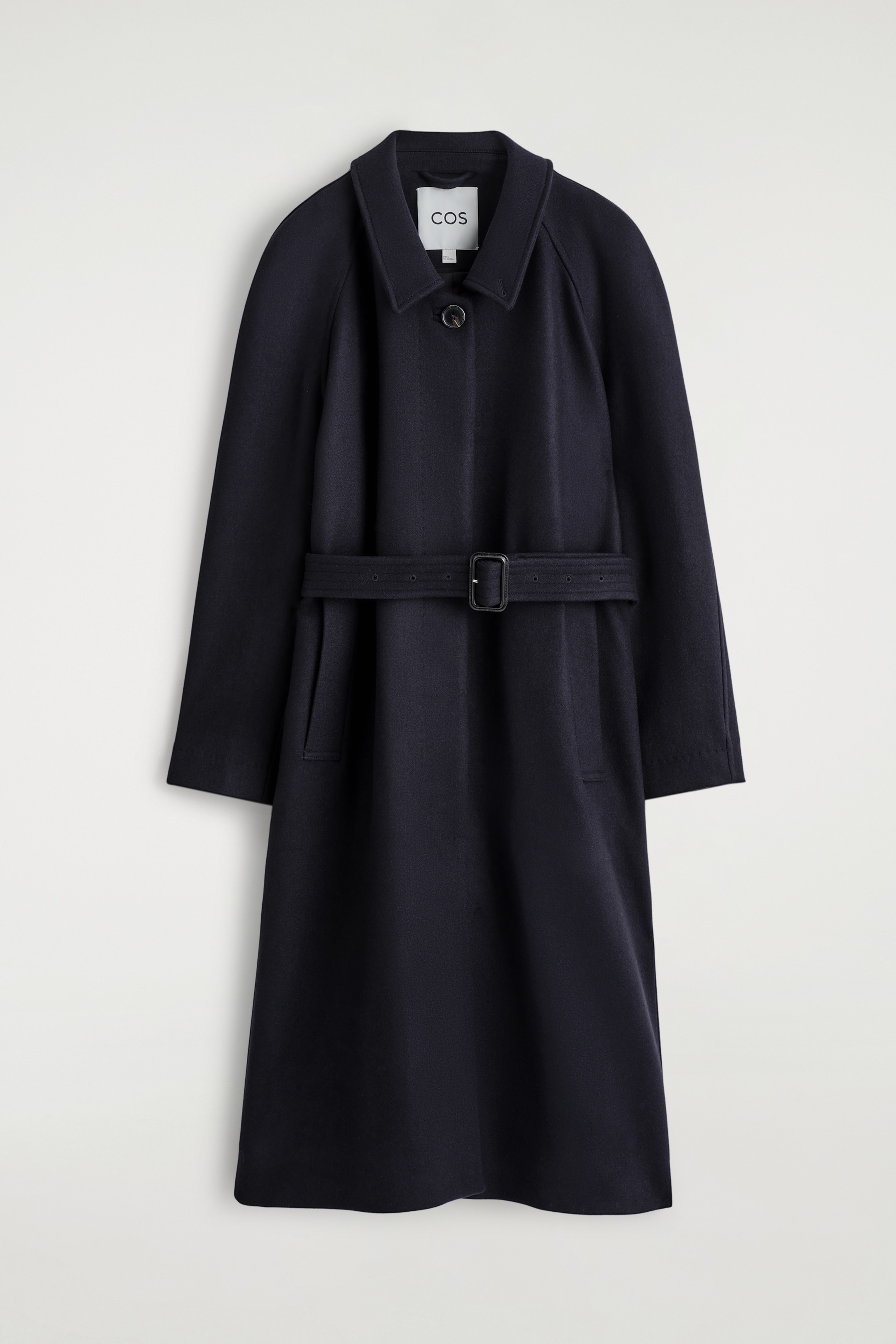 Agrandir l'image: Un manteau bleu marine foncé est présenté de face, avec une silhouette décontractée et une longueur longue. Il est conçu avec un col pointu montrant une étiquette de marque visible, des manches raglan, une fermeture à bouton unique au niveau du cou, une ceinture en tissu assorti avec une boucle rectangulaire foncée serrant la taille, et deux poches passepoilées verticales sous la ceinture.