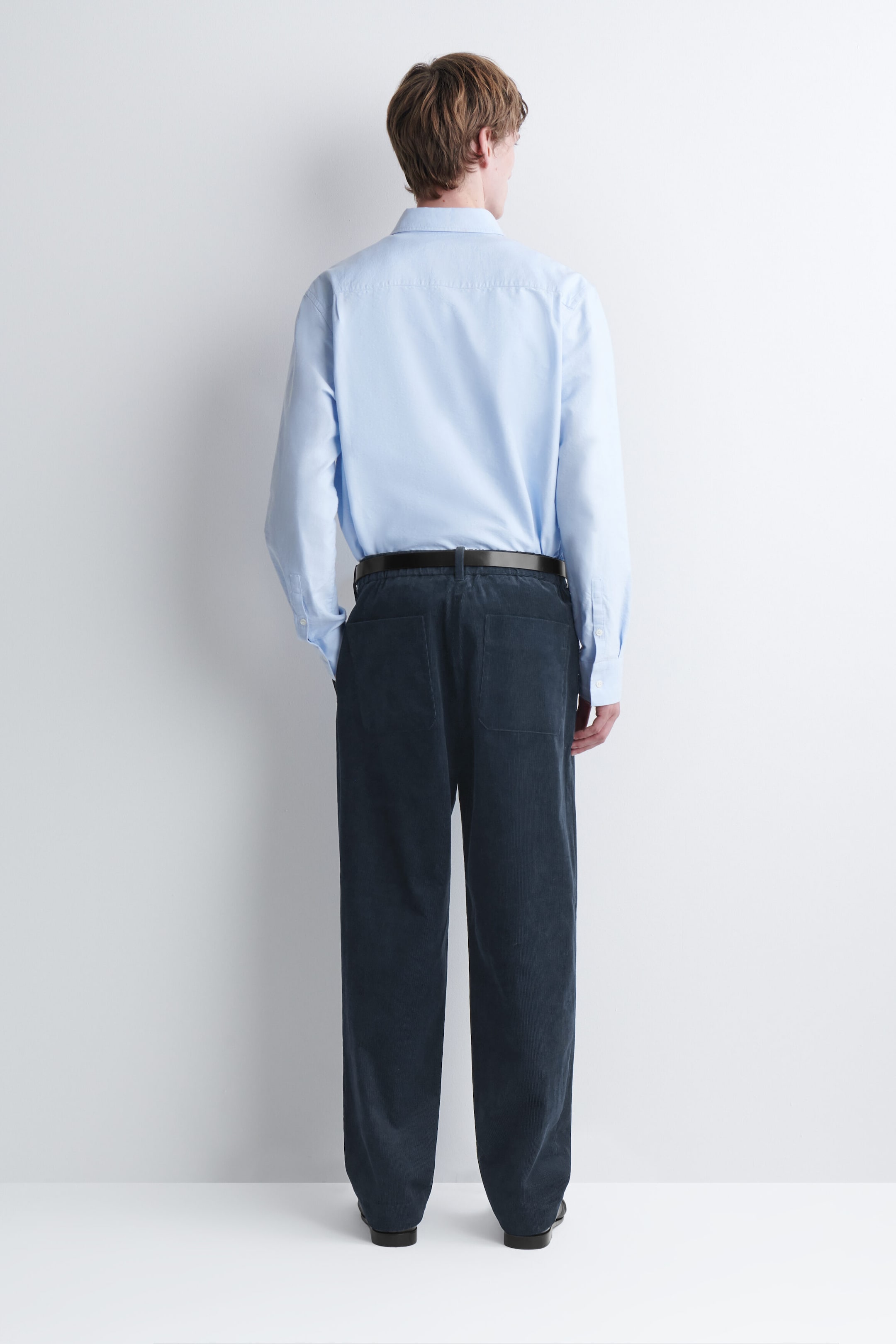 Corduroy relaxed-fit tapered pants in dark navy blue are worn by the model and shown from the back. The pants feature a relaxed fit through the leg, two back patch pockets, and are styled with a dark leather belt and a tucked-in light blue shirt. Corduroy relaxed-fit tapered pants in dark navy blue are worn by the model and shown from the back. The pants feature a relaxed fit through the leg, two back patch pockets, and are styled with a dark leather belt and a tucked-in light blue shirt.