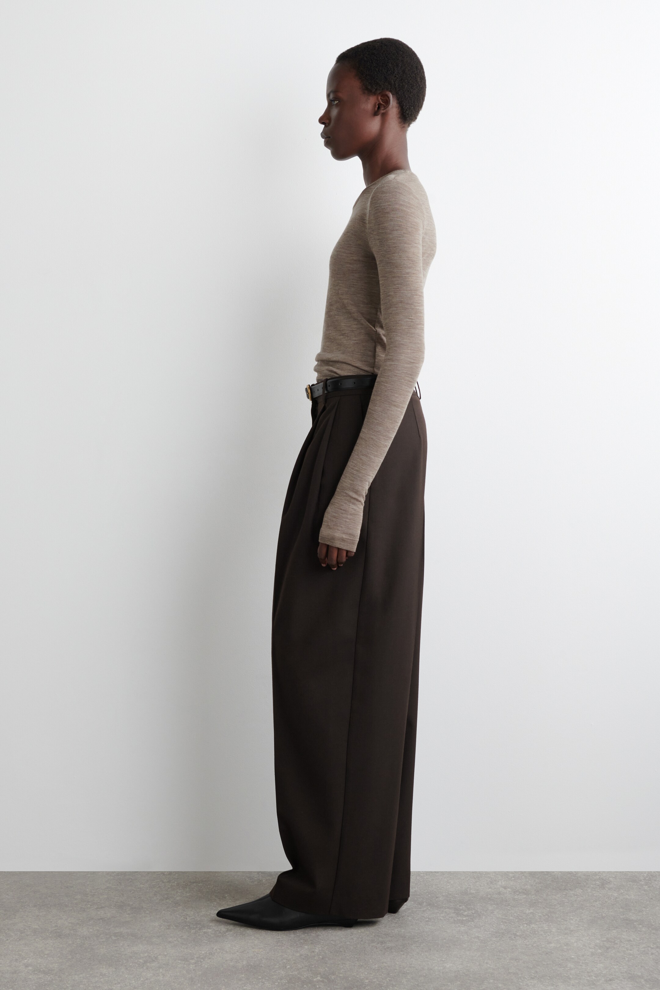 View larger image: A light brown long-sleeved top and dark brown wide-leg pleated pants are being presented in a side profile view. The top is fitting snugly, featuring long sleeves that are extending over the hands. The high-waisted pants are secured with a black belt and are falling with a full, wide leg to the floor, covering black pointed-toe flats.