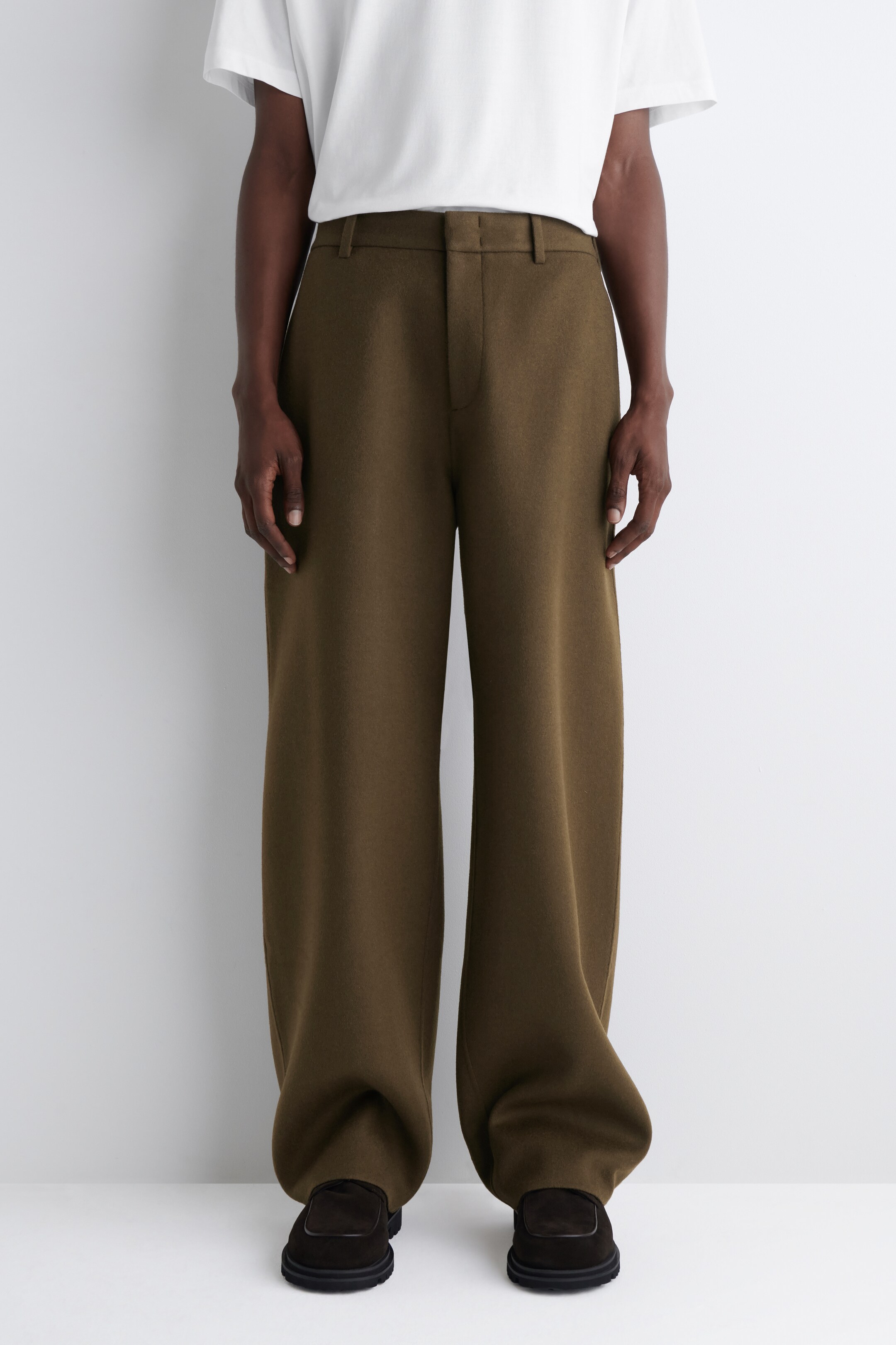 Agrandir l'image: Un pantalon vert olive à jambes larges est porté, face directement vers l'avant. Ils présentent une ceinture plate avec des passants de ceinture et une fermeture éclair dissimulée. Les jambes longues et fluides tombent largement, s'accumulant légèrement sur des chaussures sombres.