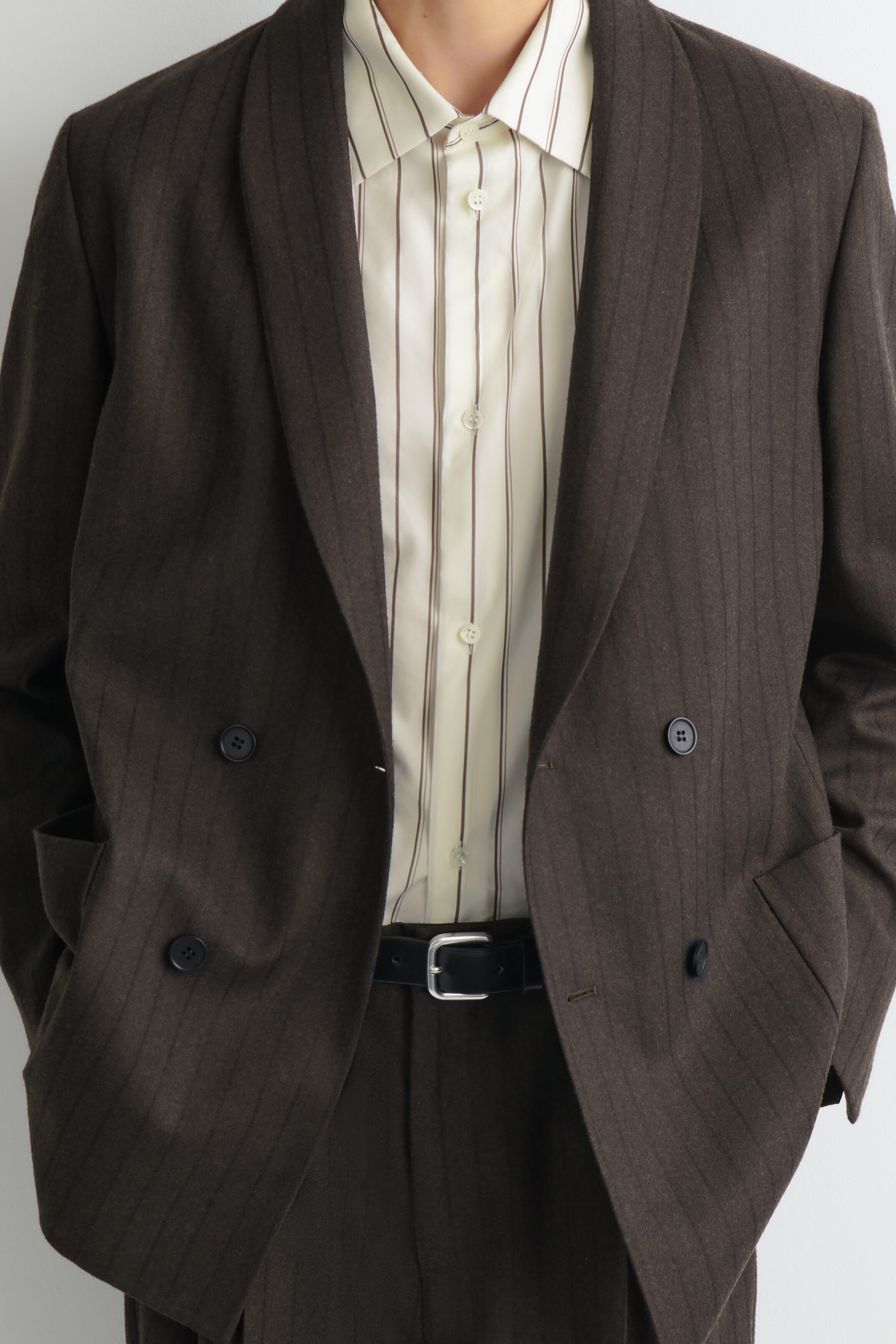A double-breasted, relaxed-fit brown pinstriped wool blazer is shown in a close-up front view, featuring shawl lapels and side pockets. The open blazer is layered over a cream-colored striped button-up shirt tucked into matching trousers, finished with a black leather belt. A double-breasted, relaxed-fit brown pinstriped wool blazer is shown in a close-up front view, featuring shawl lapels and side pockets. The open blazer is layered over a cream-colored striped button-up shirt tucked into matching trousers, finished with a black leather belt.