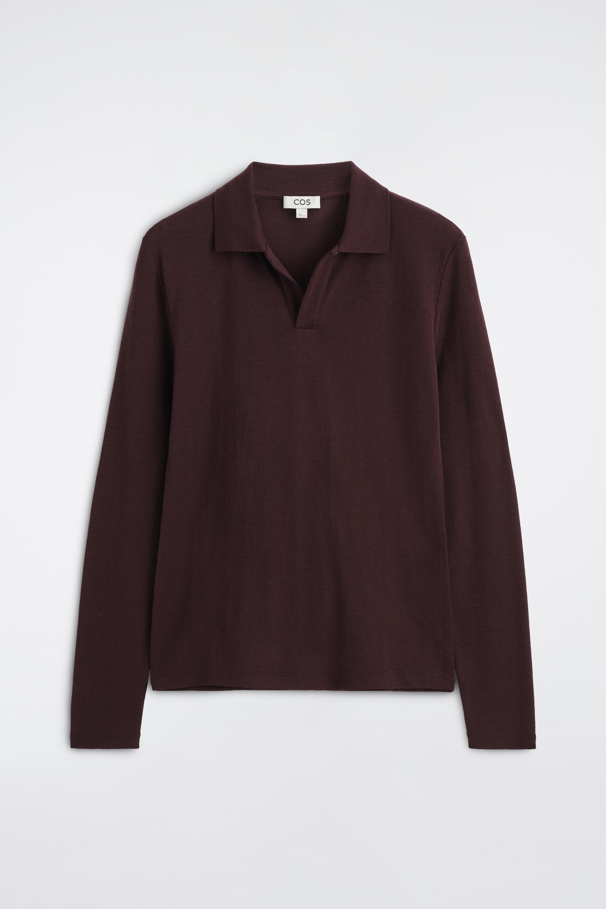 Een slank polohemd van wollen jersey met open kraag in diep bordeauxrood wordt door het model gedragen over een wit T-shirt met ronde hals. Het shirt met lange mouwen heeft een open kraag zonder knopen en is gestyled met een zwarte geplooide broek.