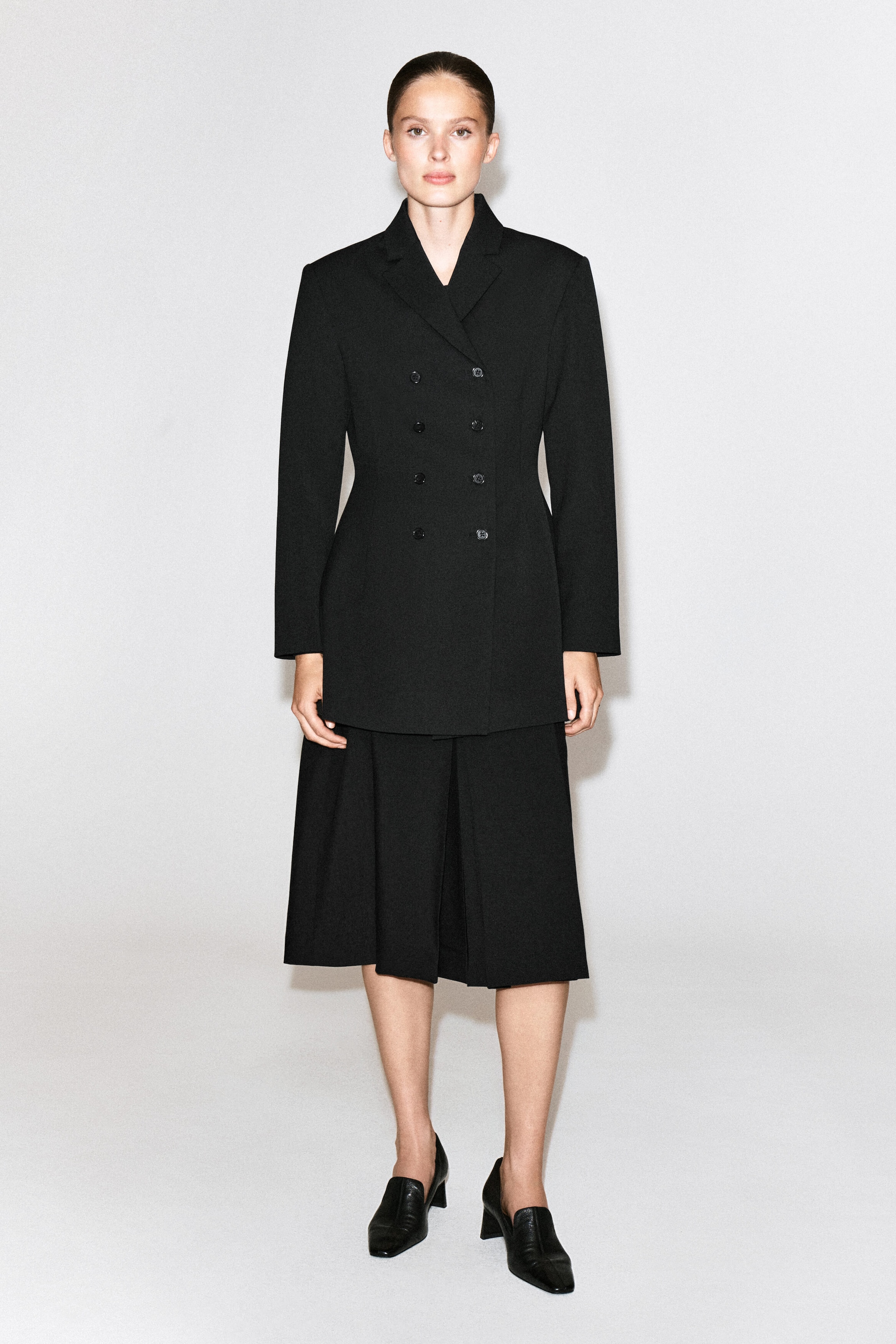 The model is wearing a black box-pleat wool midi skirt, featuring structured pleats that add volume and a mid-calf hemline. The skirt is styled with a coordinating double-breasted black blazer and black leather heeled loafers. The model is wearing a black box-pleat wool midi skirt, featuring structured pleats that add volume and a mid-calf hemline. The skirt is styled with a coordinating double-breasted black blazer and black leather heeled loafers.