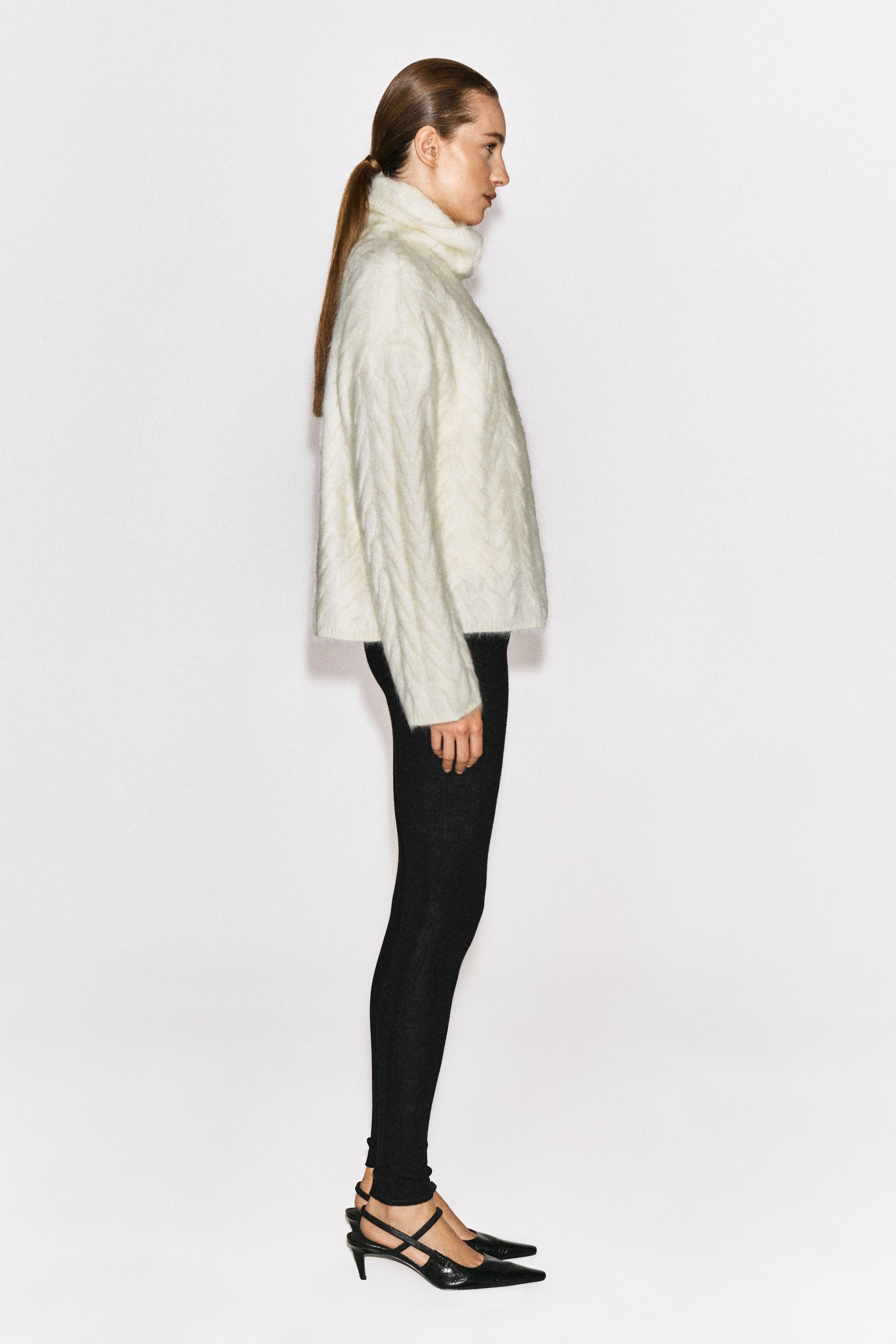 A cable-knit brushed cashmere funnel-neck jumper in off-white is worn by the model, who is standing in profile. The textured, long-sleeved jumper features a relaxed fit and is paired with black slim-fit leggings and black pointed-toe heels.