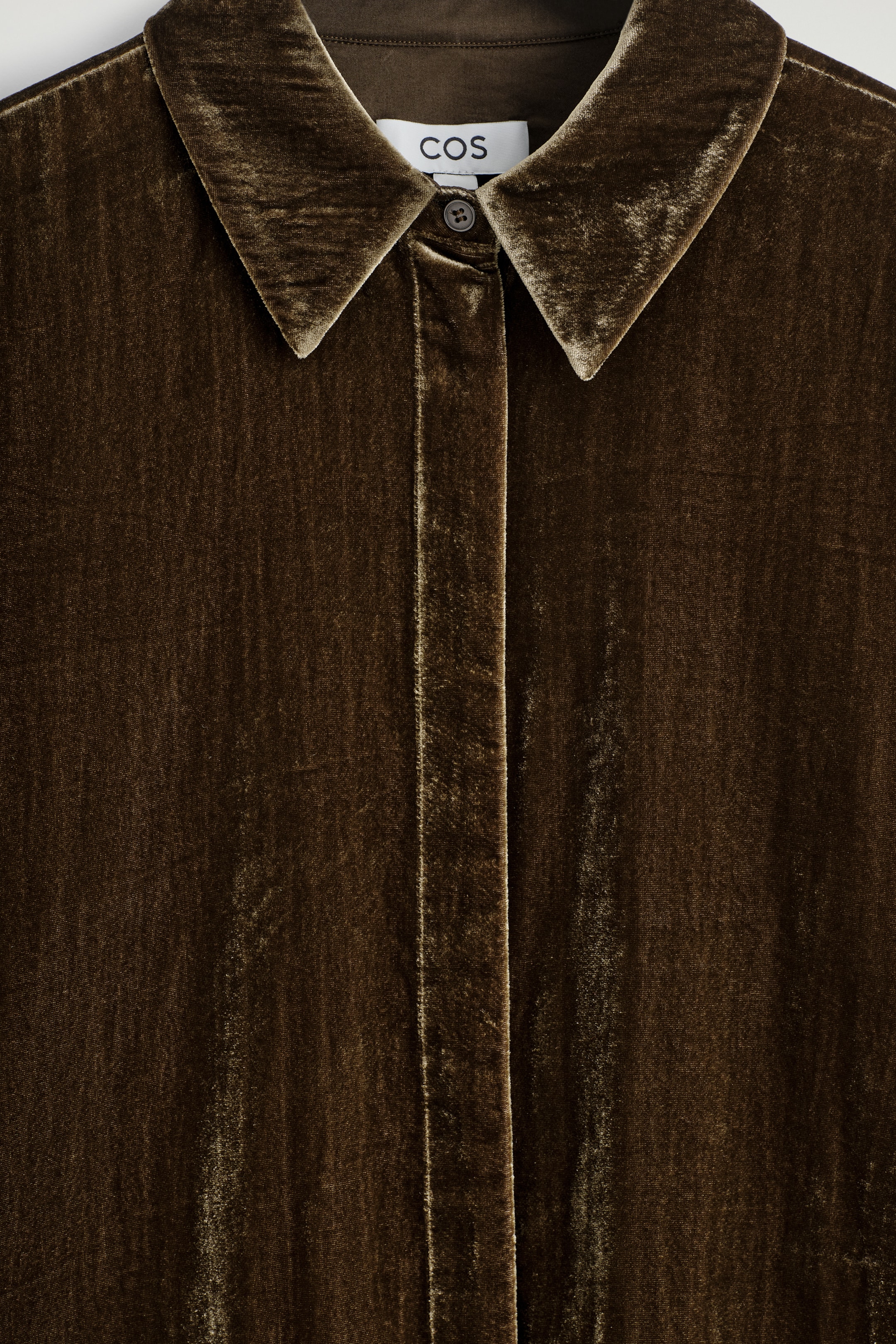 View larger image: The front of a dark brown velvet shirt is featuring a pointed collar, a button placket with one visible button at the top, and a 'COS' label. The velvet fabric is reflecting light, highlighting its tactile texture.