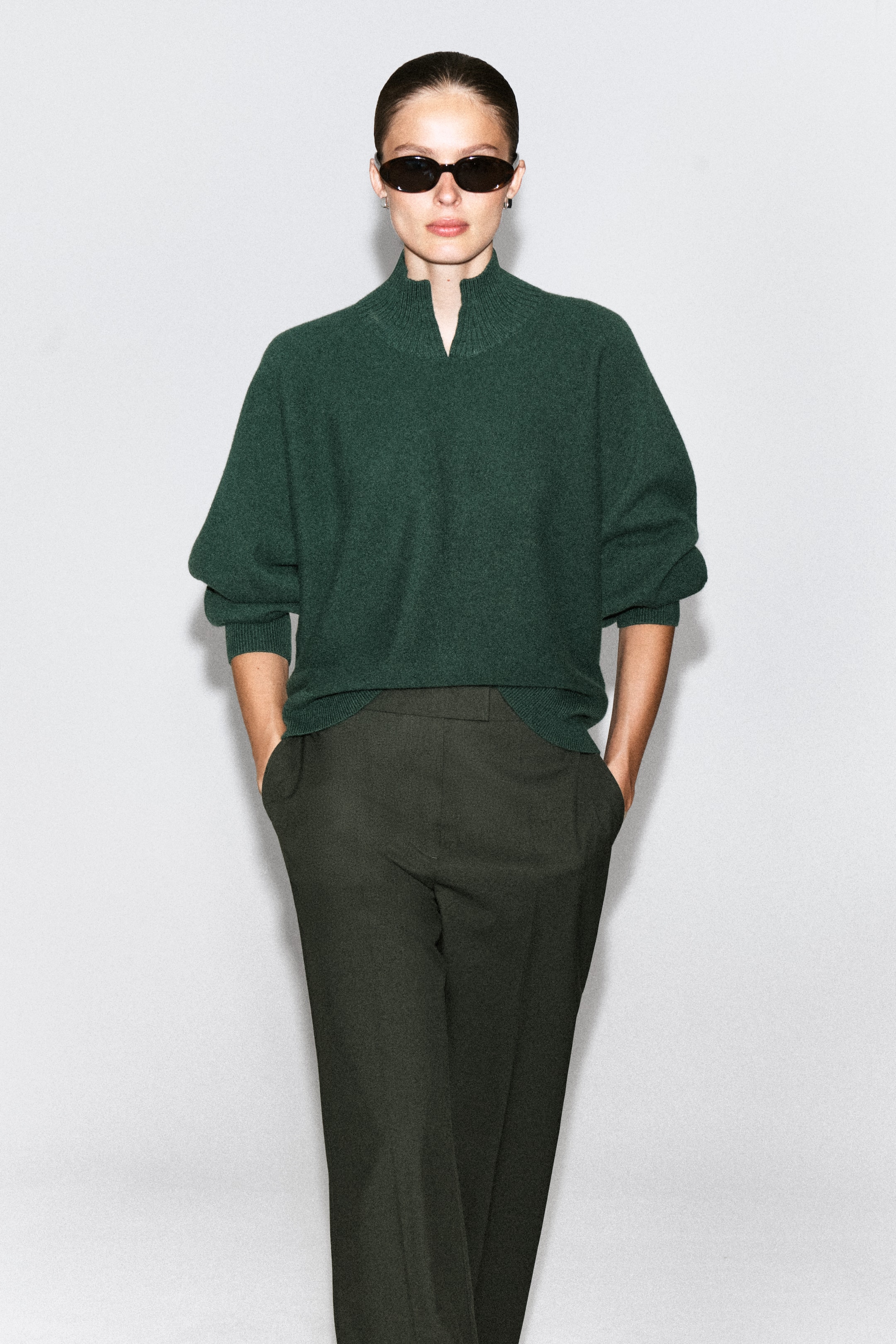 Ver imagen más grande: La modelo lleva puesto un jersey de lana afieltrada de color verde oscuro y corte holgado, con cuello alzado acanalado y detalle en V en la parte delantera. El jersey se combina con pantalones oscuros y gafas de sol negras.