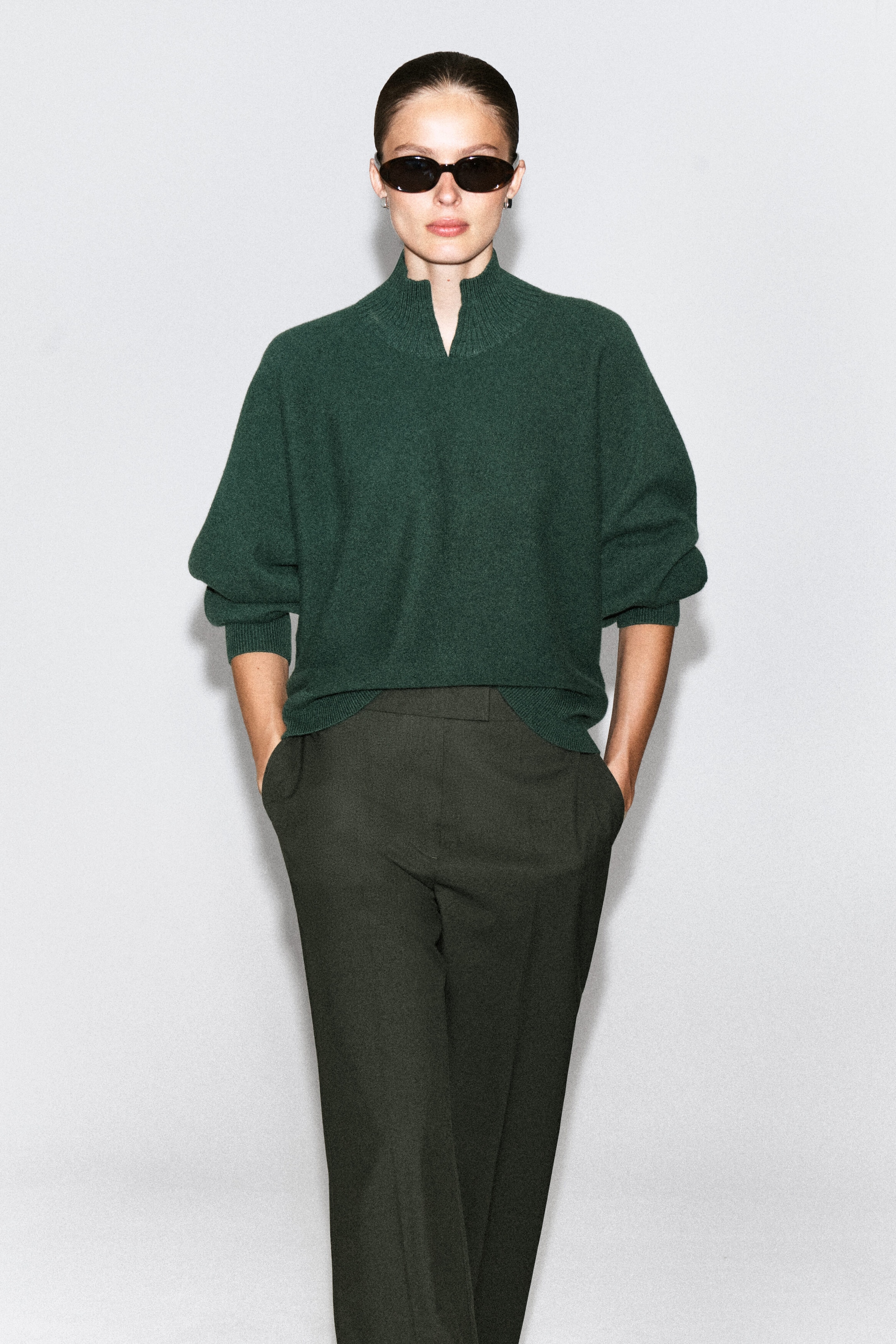 Un pull en laine bouillie vert foncé à coupe décontractée est porté par le mannequin, avec un col cheminée côtelé et un détail en V à l’avant. Le pull est porté avec un pantalon foncé et des lunettes de soleil noires. Un pull en laine bouillie vert foncé à coupe décontractée est porté par le mannequin, avec un col cheminée côtelé et un détail en V à l’avant. Le pull est porté avec un pantalon foncé et des lunettes de soleil noires.