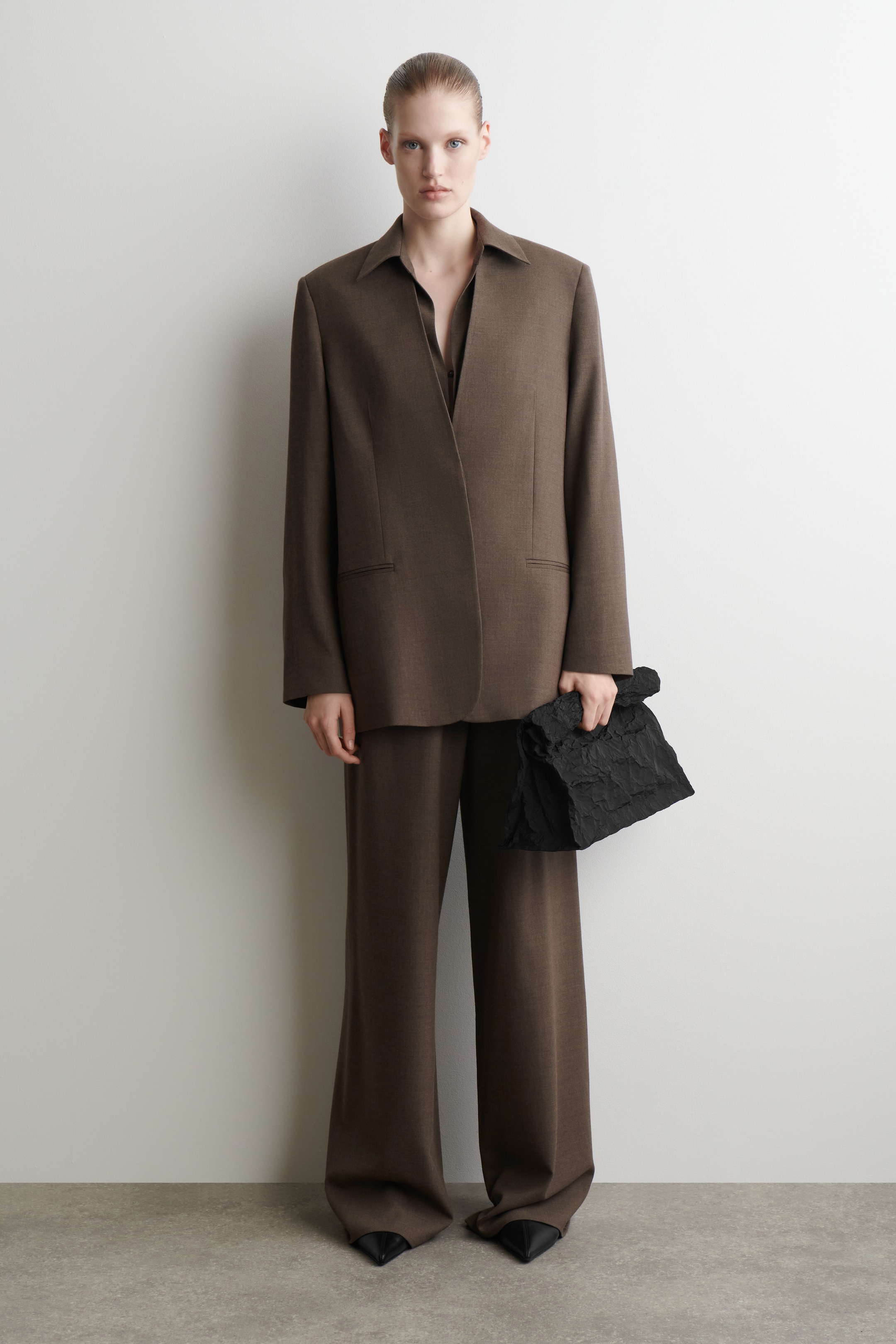 Visualizza immagine più grande: Un abito marrone viene presentato frontalmente, caratterizzato da un blazer oversize con colletto a punta e due tasche con patta, stratificato su una camicia scura. I pantaloni a gamba larga abbinati completano la silhouette rilassata, mentre una borsa a mano nera con texture viene tenuta in mano e sono visibili scarpe nere a punta.