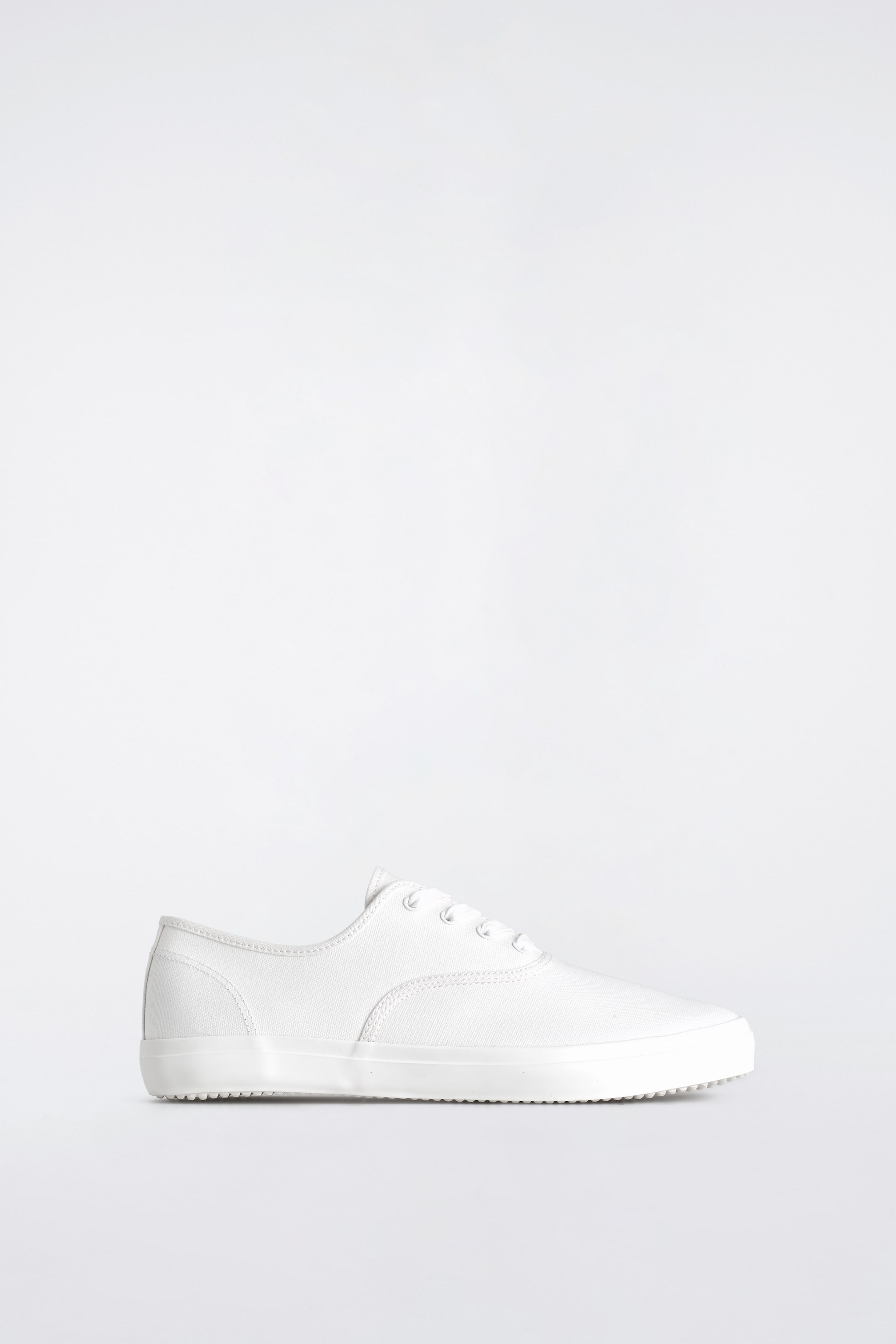 Visualizza immagine più grande: Una sneaker bianca in tela a taglio basso è mostrata dal lato destro, leggermente inclinata in avanti. Presenta lacci bianchi infilati attraverso occhielli, una punta arrotondata e una suola piatta in gomma bianca con una parete laterale testurizzata.