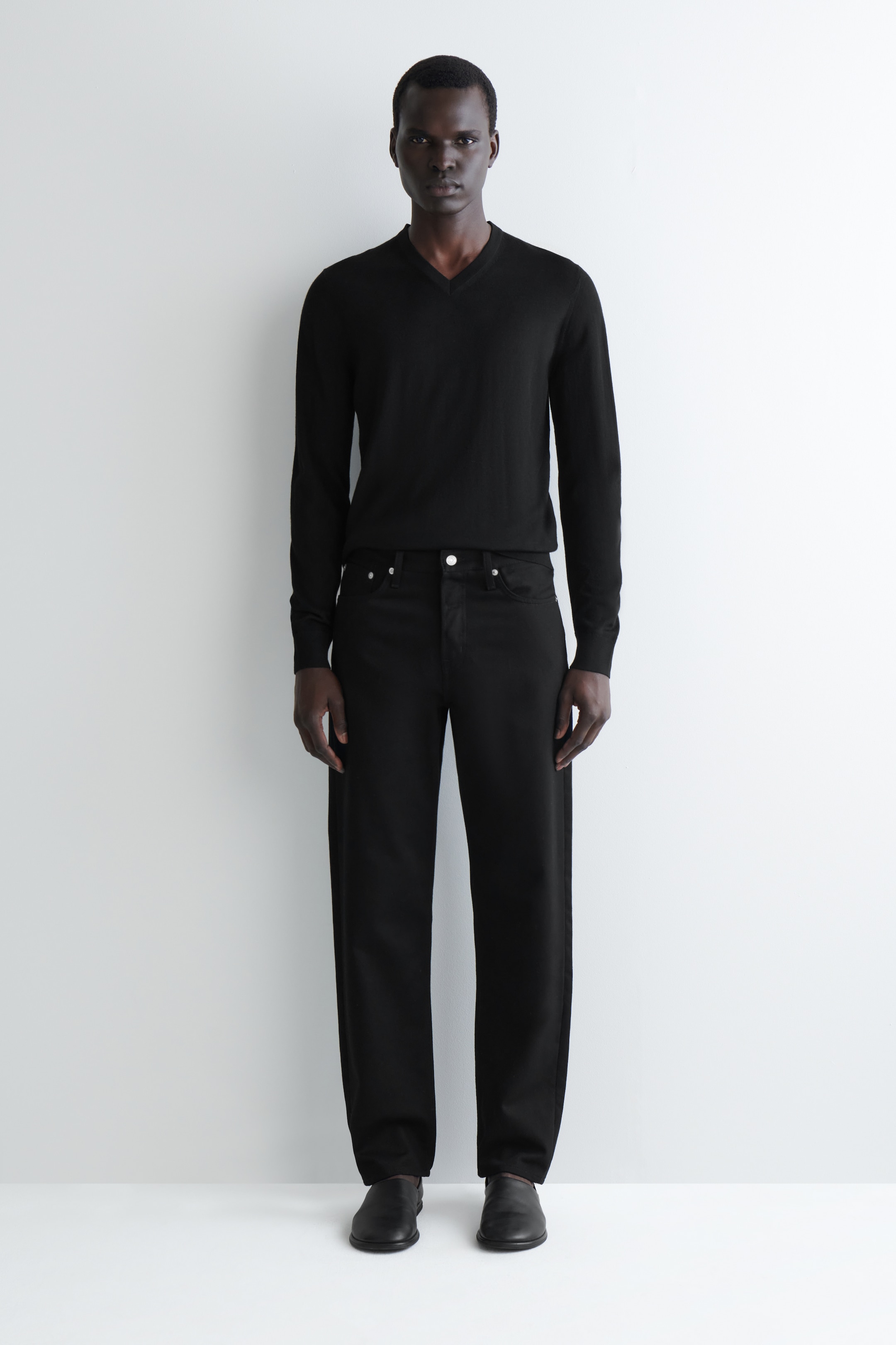 Visualizza immagine più grande: Un maglione nero con scollo a V viene presentato, caratterizzato da maniche lunghe e un orlo dritto. Vengono mostrati anche jeans neri a vestibilità rilassata, con gamba dritta, rivetti d'argento visibili e un bottone frontale. Scarpe nere slip-on completano il look.