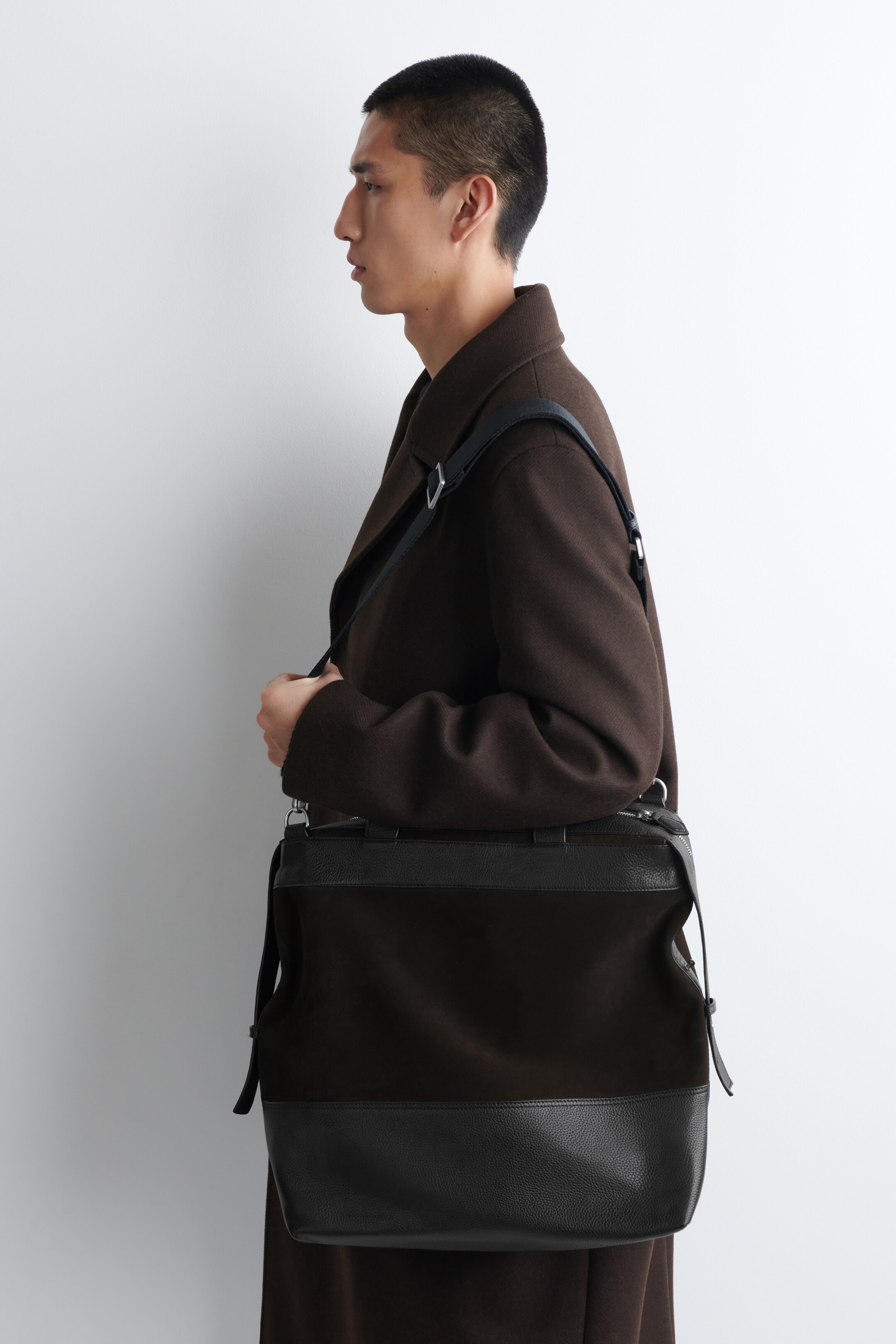 Visualizza immagine più grande: Una borsa a tracolla nera è indossata trasversalmente sul corpo, leggermente inclinata verso sinistra, sopra un cappotto marrone scuro. La borsa presenta un design bicolore con un tessuto scuro e liscio nella sua sezione superiore e una pelle nera testurizzata e ciottolata nella sua sezione inferiore, mentre una cerniera superiore, piccoli manici per il trasporto e una tracolla nera con ferramenta color argento sono anche visibili.