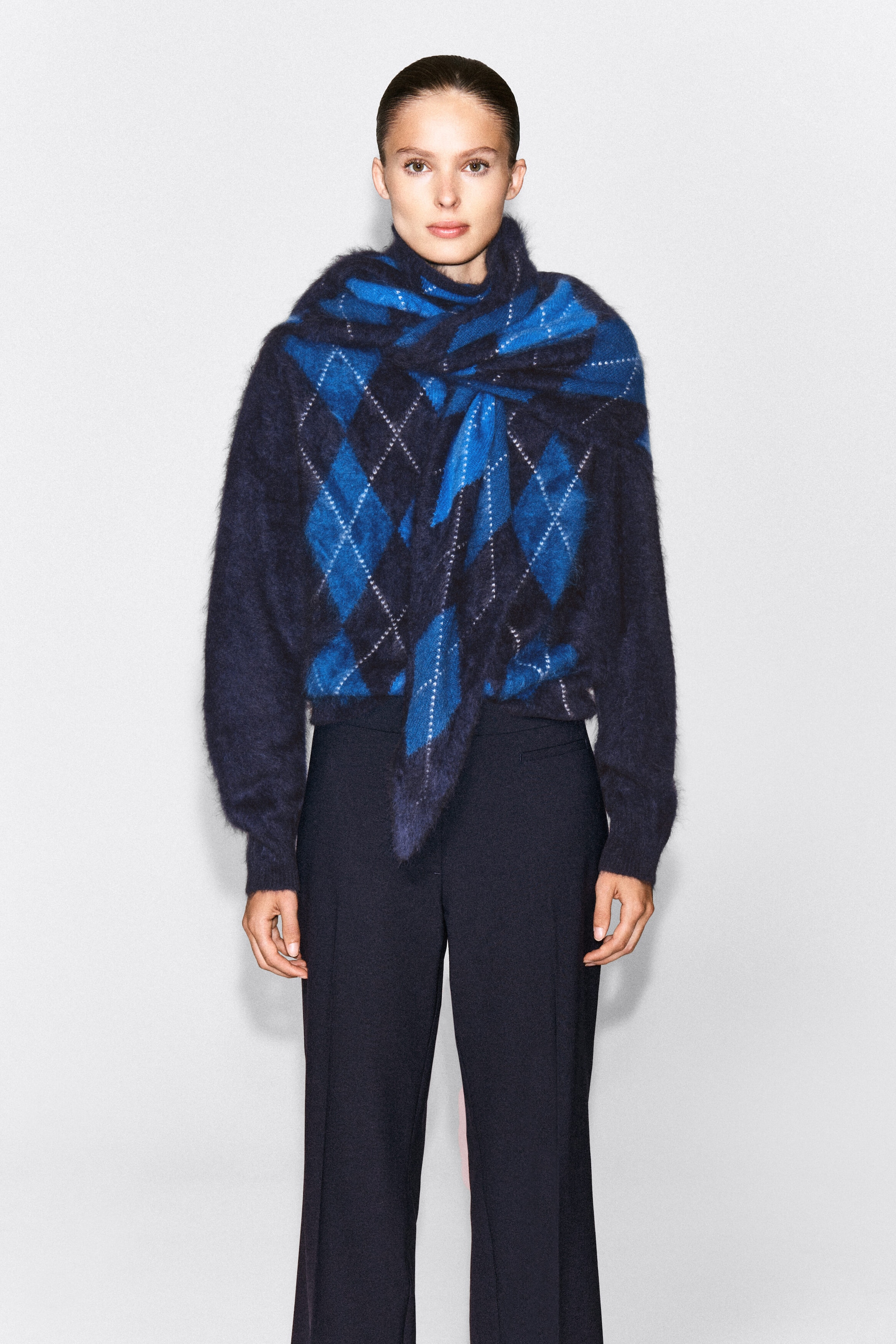 An argyle brushed cashmere triangle scarf in navy and bright blue is worn by the model, draped over the shoulders and tied loosely at the front. The scarf is layered over a matching argyle knit jumper and paired with dark trousers, highlighting its soft, fuzzy texture. An argyle brushed cashmere triangle scarf in navy and bright blue is worn by the model, draped over the shoulders and tied loosely at the front. The scarf is layered over a matching argyle knit jumper and paired with dark trousers, highlighting its soft, fuzzy texture.
