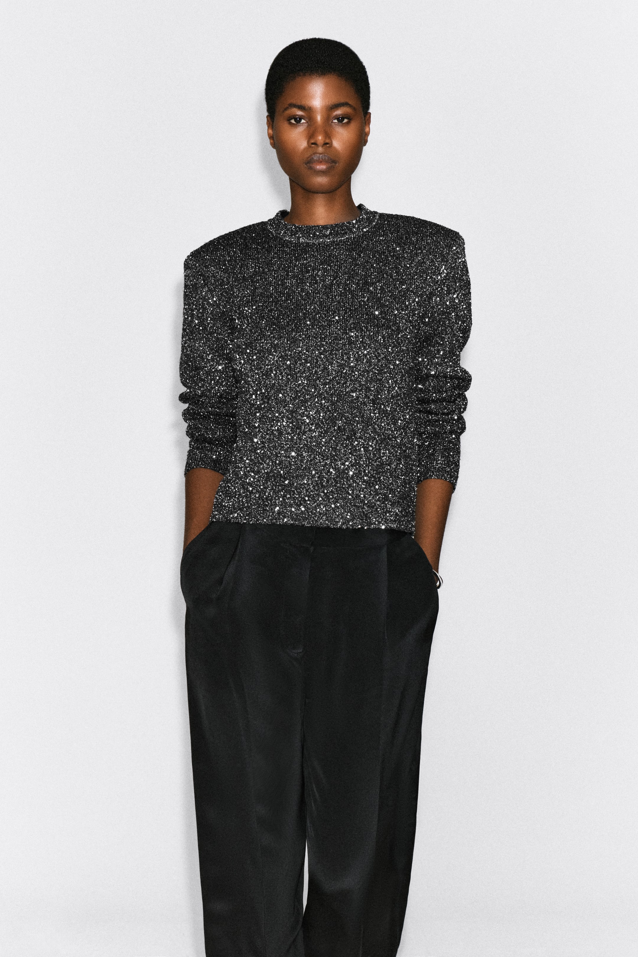 View larger image: A dark grey crew-neck jumper with a textured bouclé finish and interwoven silver sequins is being worn by the model. The jumper features long sleeves and structured shoulders and is styled with black trousers.