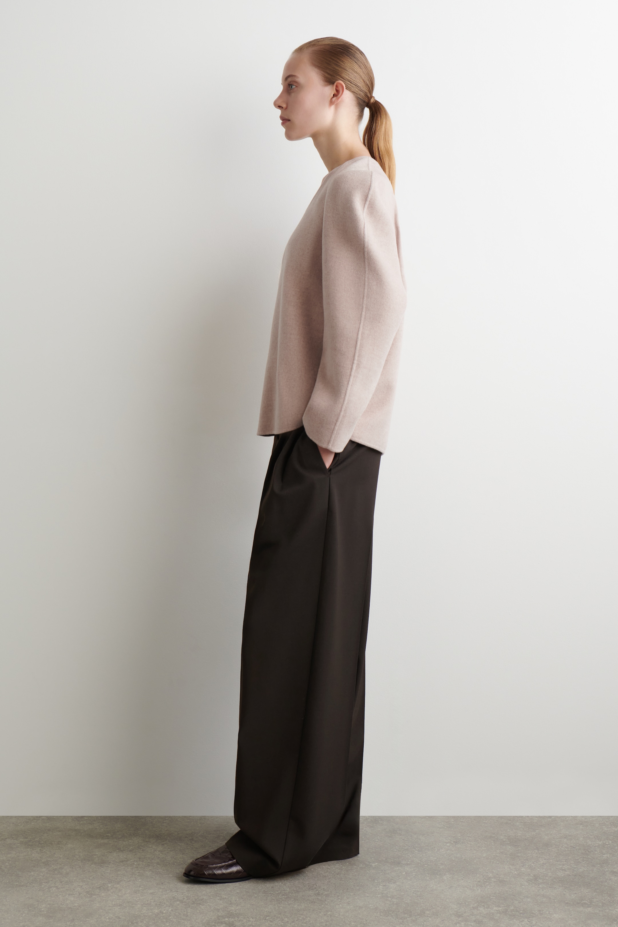 View larger image: The product, a pale blush knit top and dark brown wide-leg pants, is being presented from a side profile. The relaxed-fit top features a crew neck and drop shoulders, while the long, flowing pants are partially obscuring a dark brown textured loafer, with a hand being placed in a side pocket.