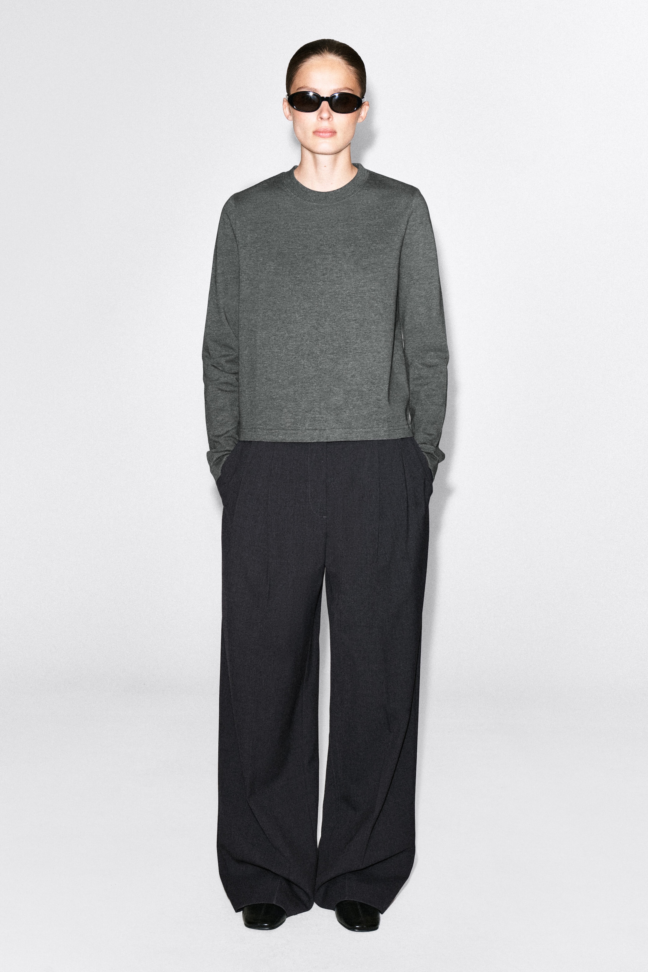 A gray, long-sleeved T-shirt featuring a crew neck and a straight hem is being worn by the model. The garment is paired with dark, wide-leg trousers and black sunglasses.