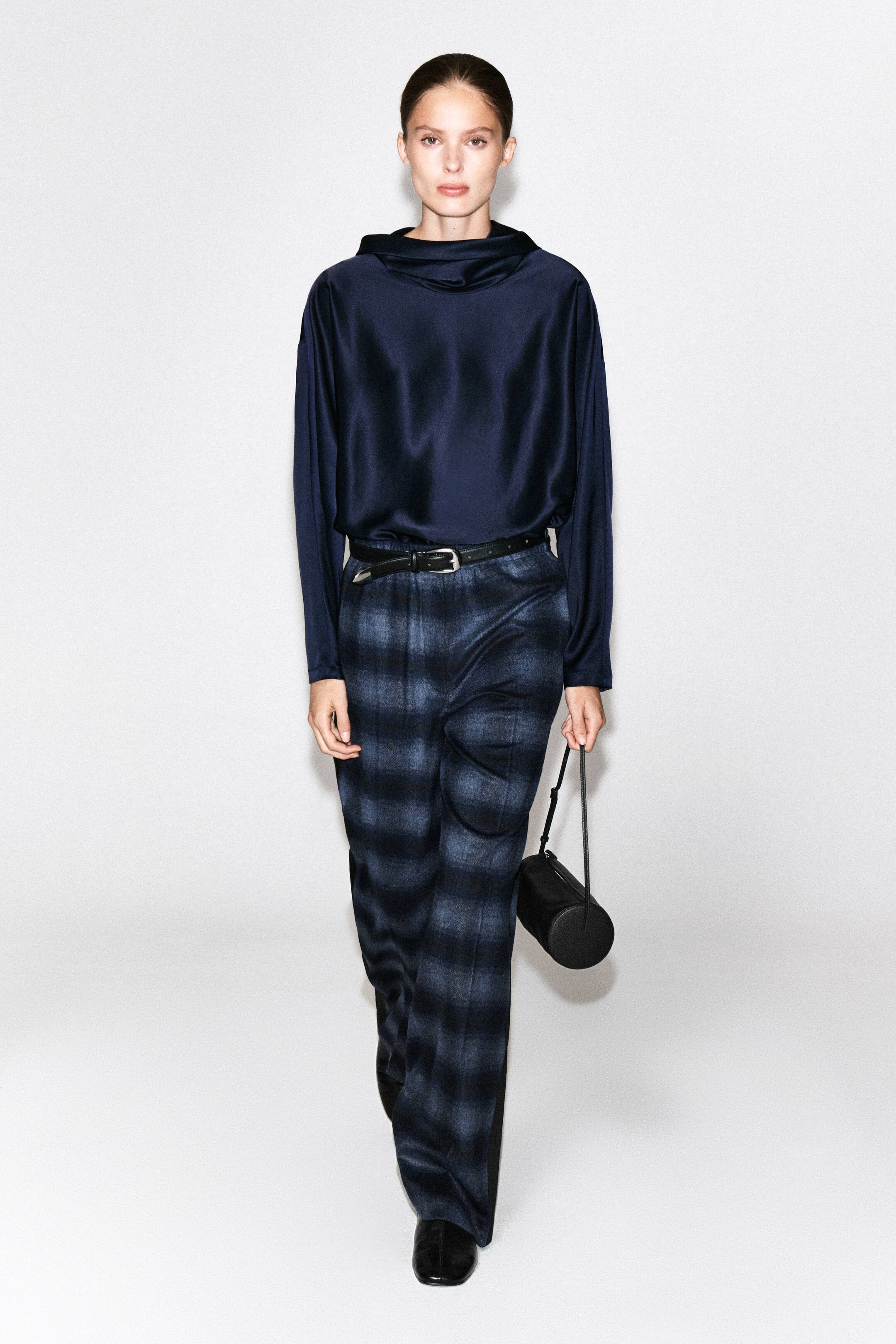 Agrandir l'image: Le mannequin porte un pantalon en mélange de laine à carreaux, présentant un motif tartan bleu foncé et noir, avec une bande latérale en satin noir. Le pantalon est associé à un haut en satin bleu marine à manches longues, rentré à l’intérieur, une fine ceinture en cuir noir et des chaussures en cuir noir.