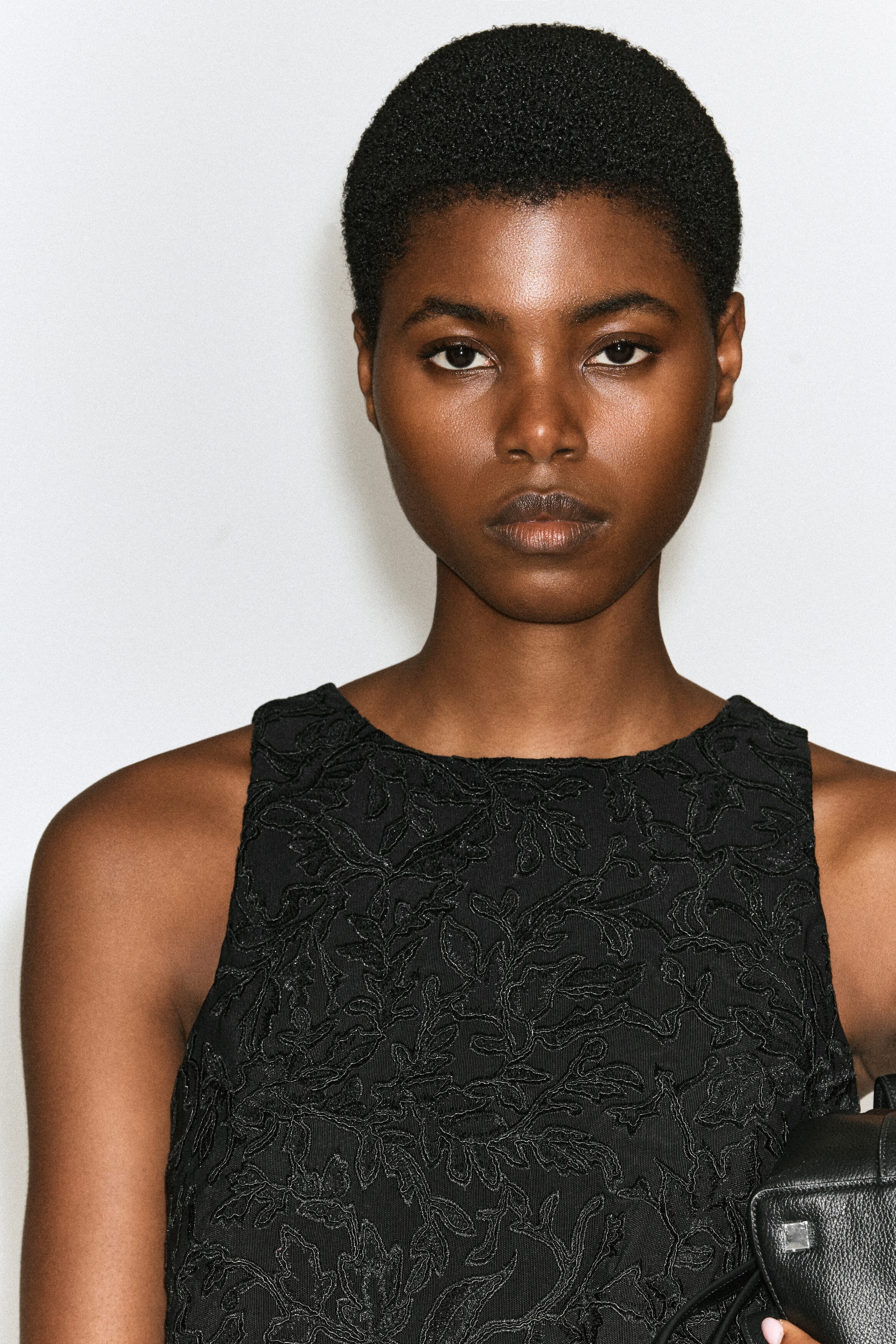 Un haut noir sans manches mettant en valeur une broderie florale complexe et un travail de dentelle texturée est porté par le mannequin. Le haut est conçu avec un col rond montant.