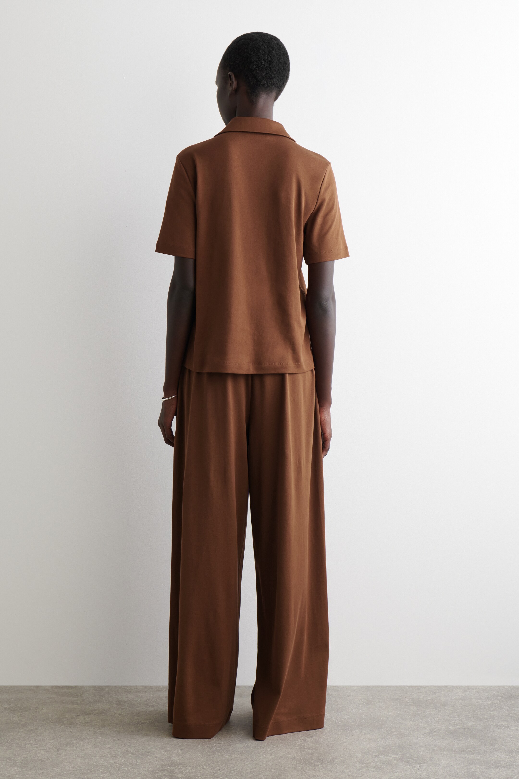 Agrandir l'image: Un ensemble deux pièces marron est présenté de dos, montrant un haut à manches courtes avec un col évasé et un pantalon à jambes larges. Le haut présente un ourlet droit, et le pantalon arbore une silhouette fluide et longue dans un tissu assorti.