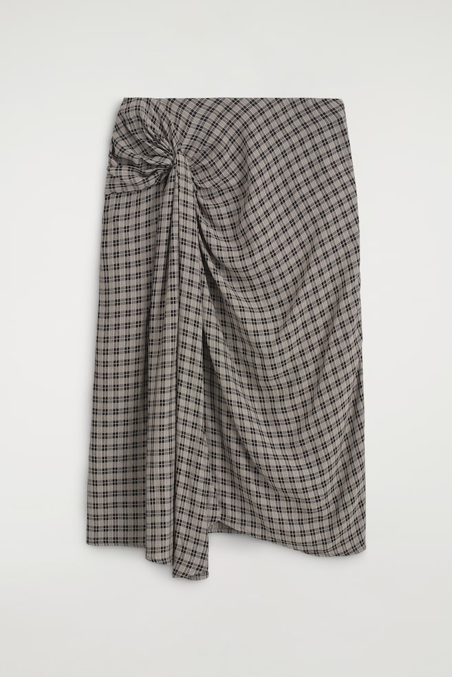 A draped checked midi skirt is shown from the front, featuring a black and beige plaid pattern. The skirt displays a gathered twist detail at the side, creating cascading folds across the front and an asymmetrical hemline. - 1