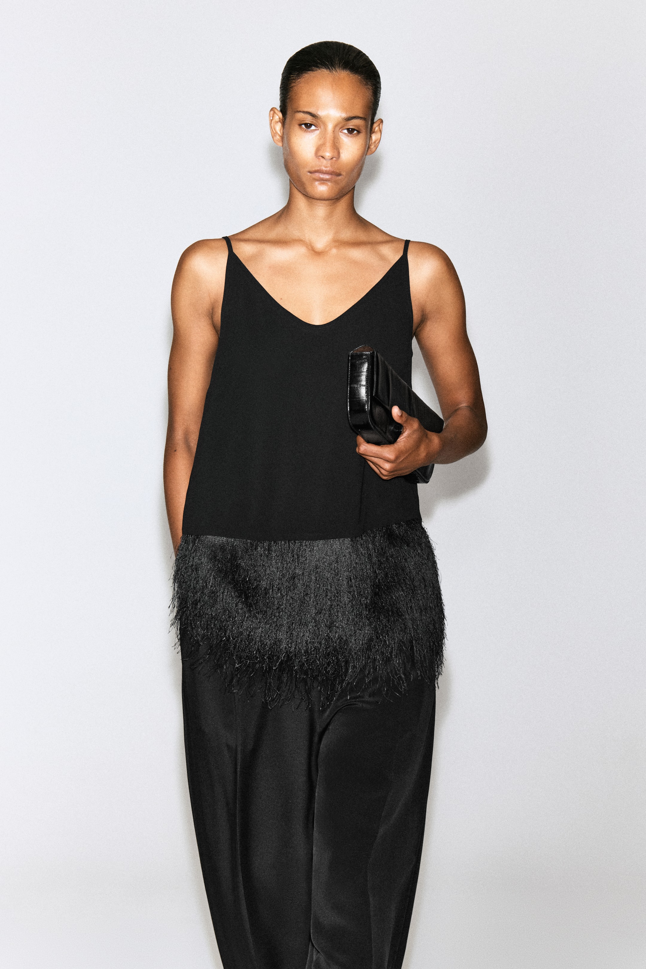 View larger image: A black cami top featuring a deep V-neckline, thin shoulder straps and a wide, textured fringe hem is worn by the model. The top is styled with black trousers, and the model is holding a black leather clutch.
