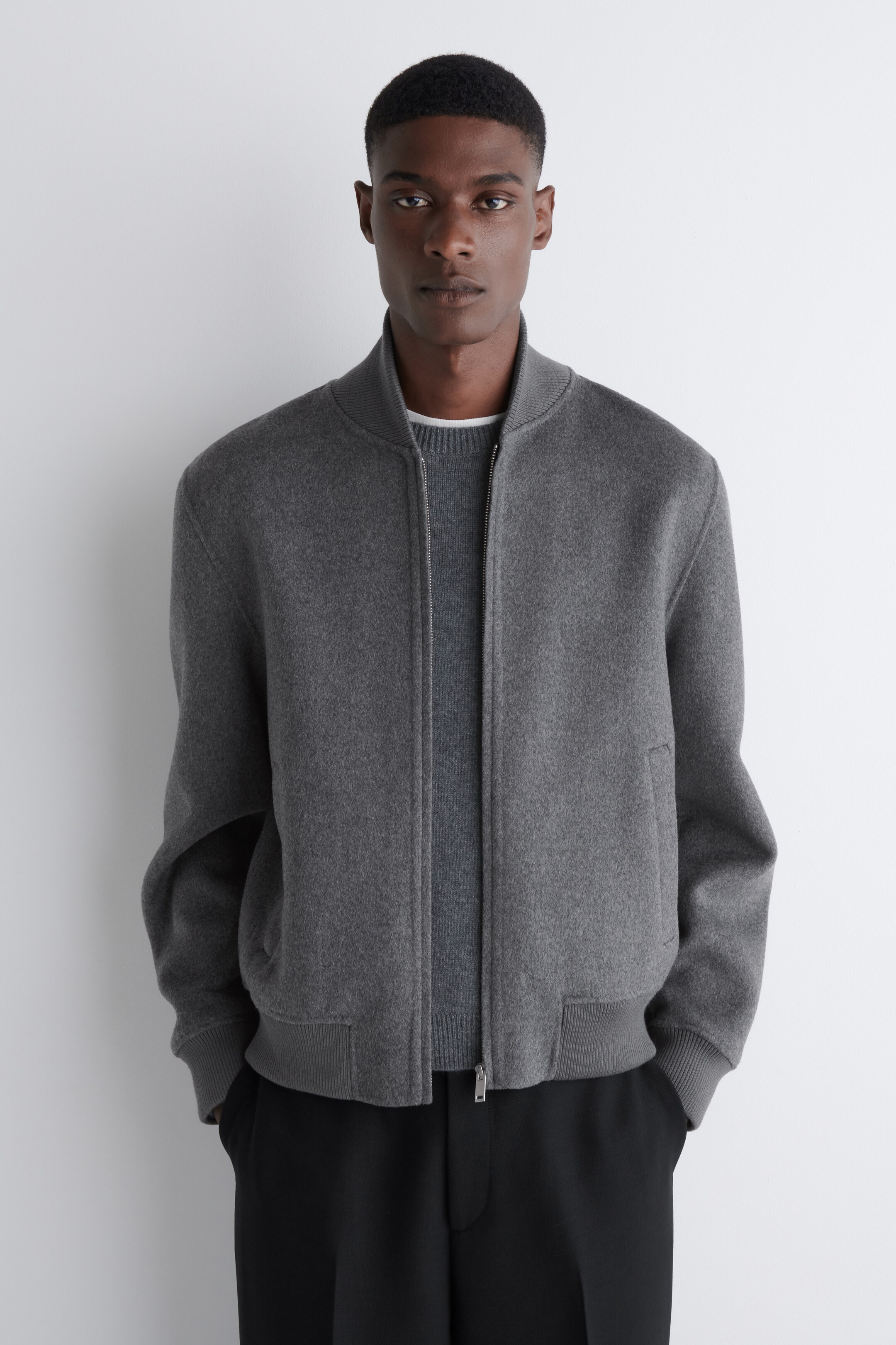 View larger image: A dark gray bomber jacket is being worn, featuring a full-length zipper closure, a ribbed stand-up collar, ribbed cuffs, and a ribbed hem. Two angled welt pockets are visible on the sides, and the jacket is layered over a lighter gray crewneck sweater.