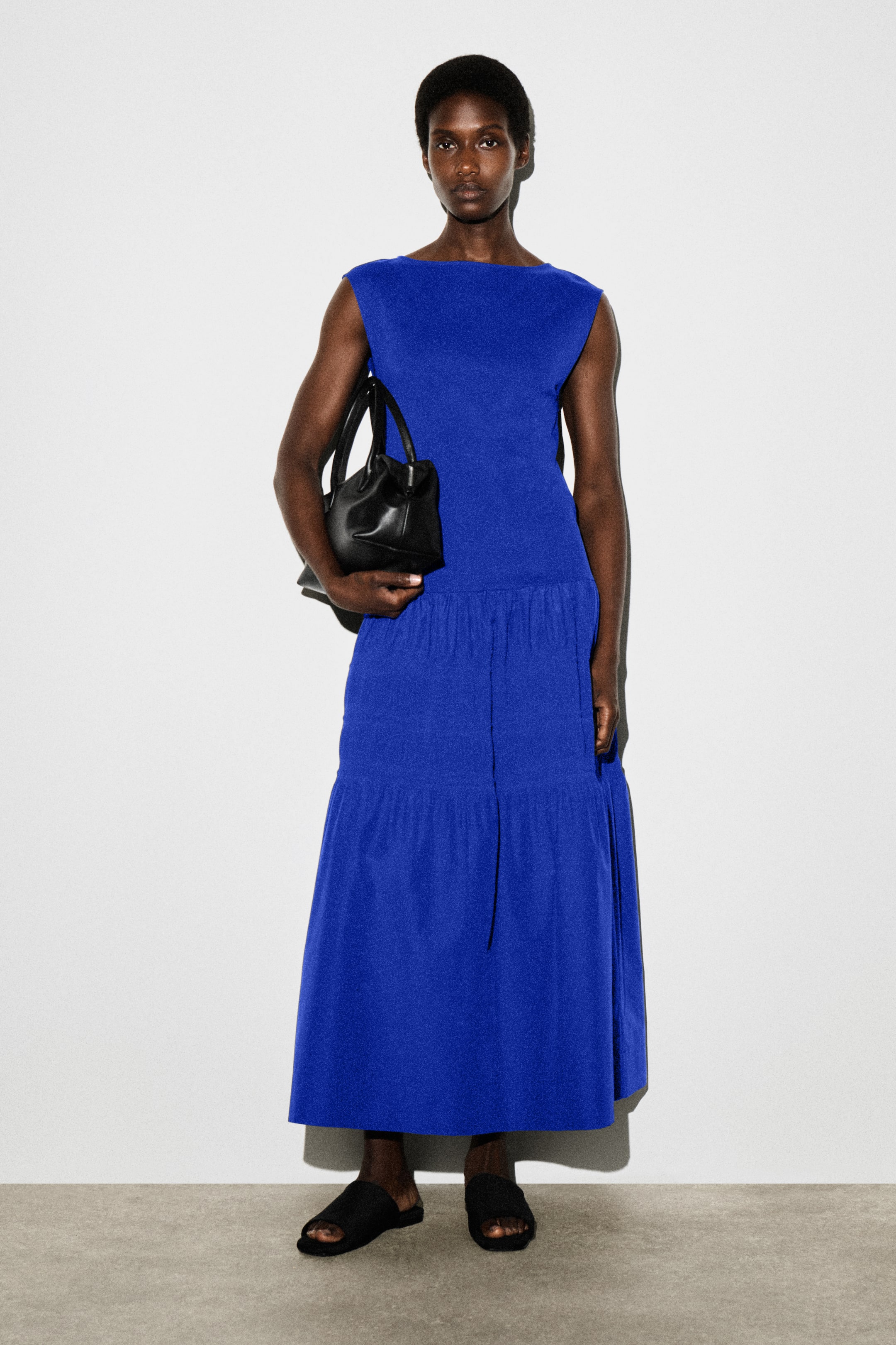 Grösseres Bild ansehen: Ein leuchtend blaues Midikleid wird vom Model getragen. Das Kleid hat ein ärmelloses Jersey-Oberteil und einen kontrastierenden, gewebten Stufenrock mit gerafften Details. Das Model hält eine schwarze Handtasche und trägt schwarze Pantoletten.