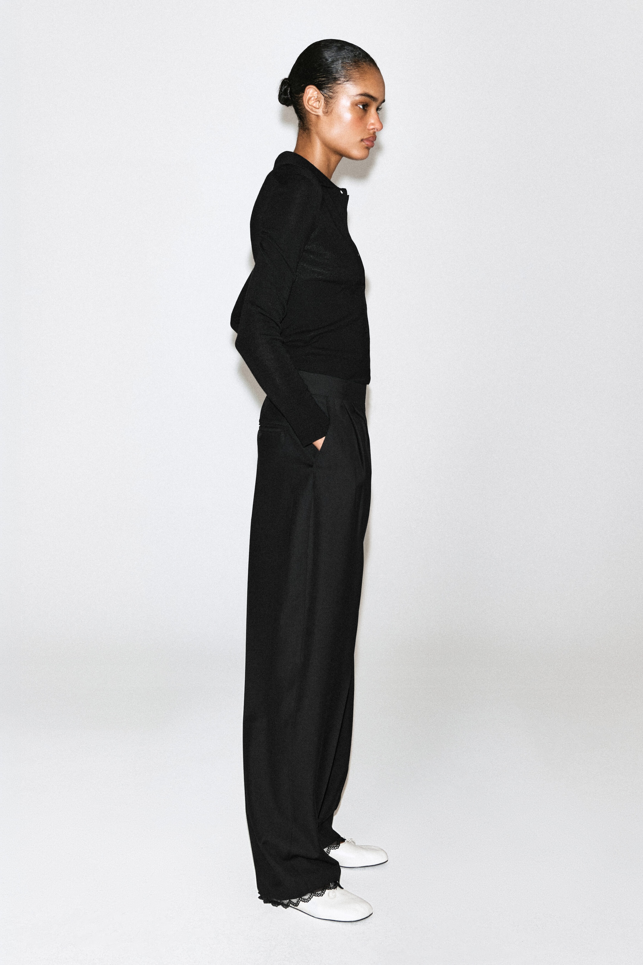 These black wide-leg trousers are shown from a side profile facing right, featuring a high-waisted fit with side slash pockets and a relaxed silhouette that drapes down to a delicate black lace trim at the hem. These black wide-leg trousers are shown from a side profile facing right, featuring a high-waisted fit with side slash pockets and a relaxed silhouette that drapes down to a delicate black lace trim at the hem.