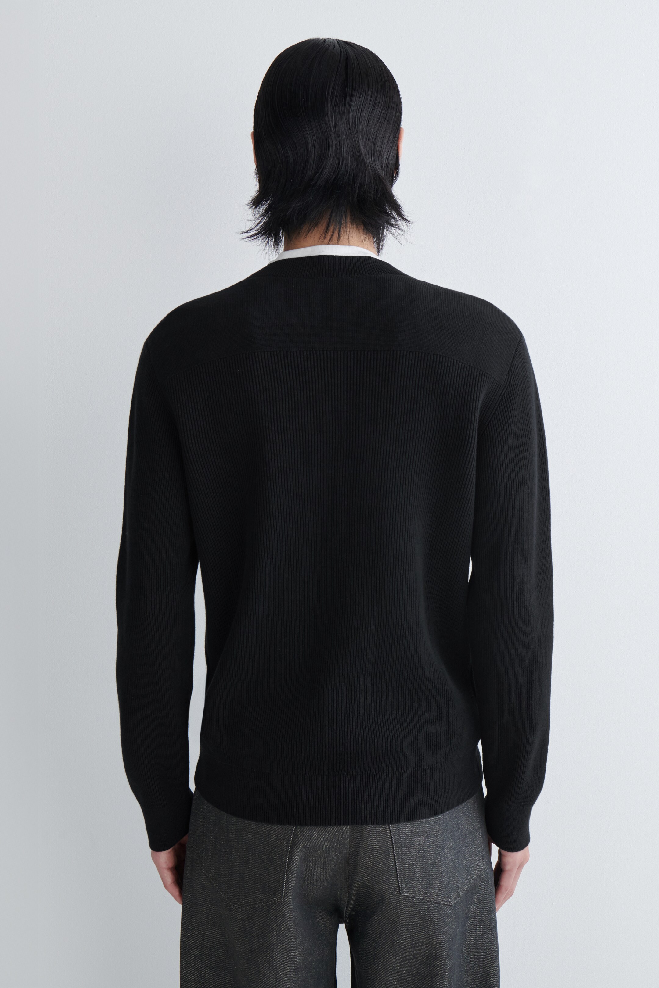 Agrandir l'image: Un pull noir côtelé est porté, vu de dos. Il présente un panneau lisse et horizontal sur le haut du dos, contrastant avec le côtelage vertical du corps principal, et un col blanc est visible dépassant à l'encolure.