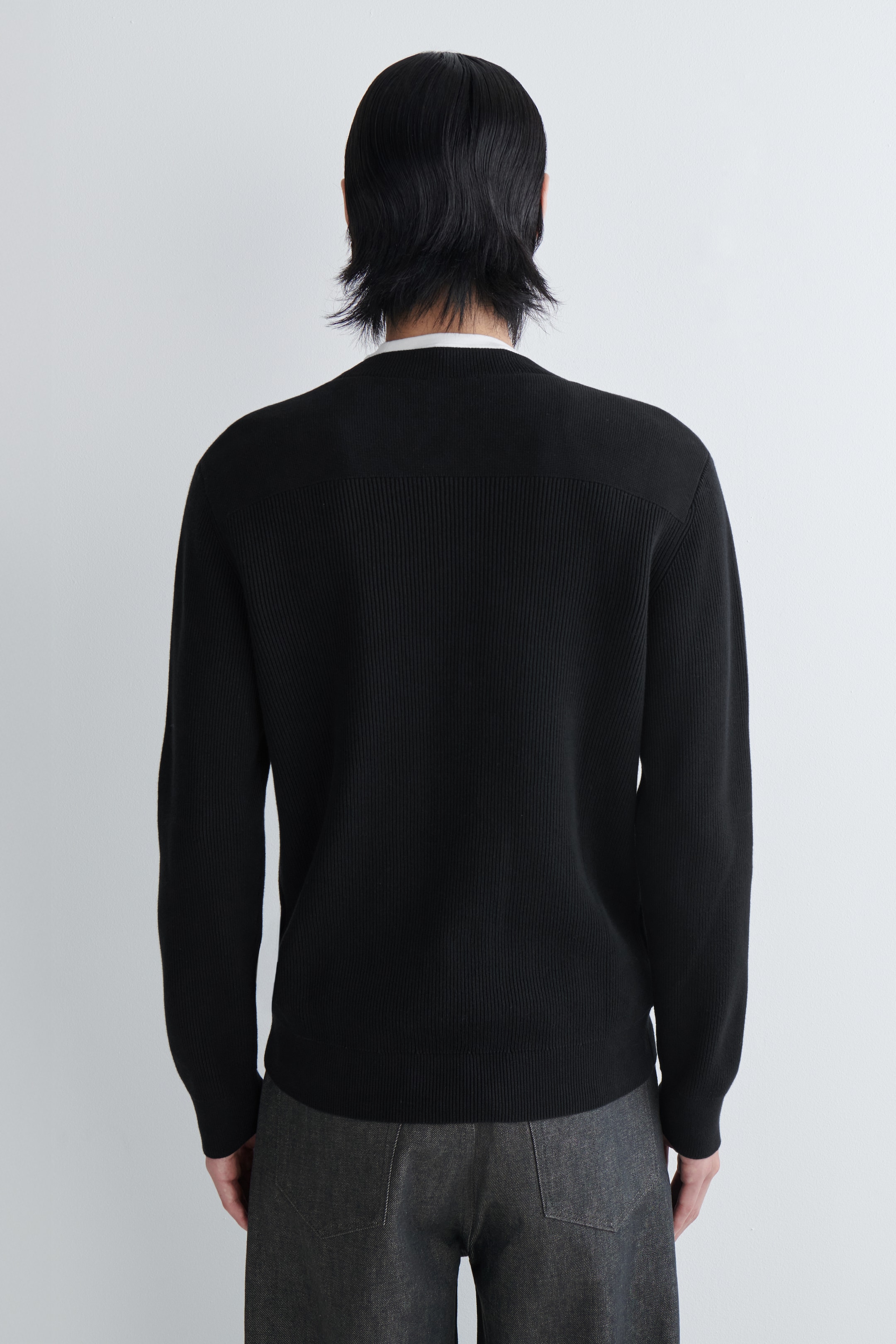 Agrandir l'image: Un pull noir côtelé est porté, vu de dos. Il présente un panneau lisse et horizontal sur le haut du dos, contrastant avec le côtelage vertical du corps principal, et un col blanc est visible dépassant à l'encolure.