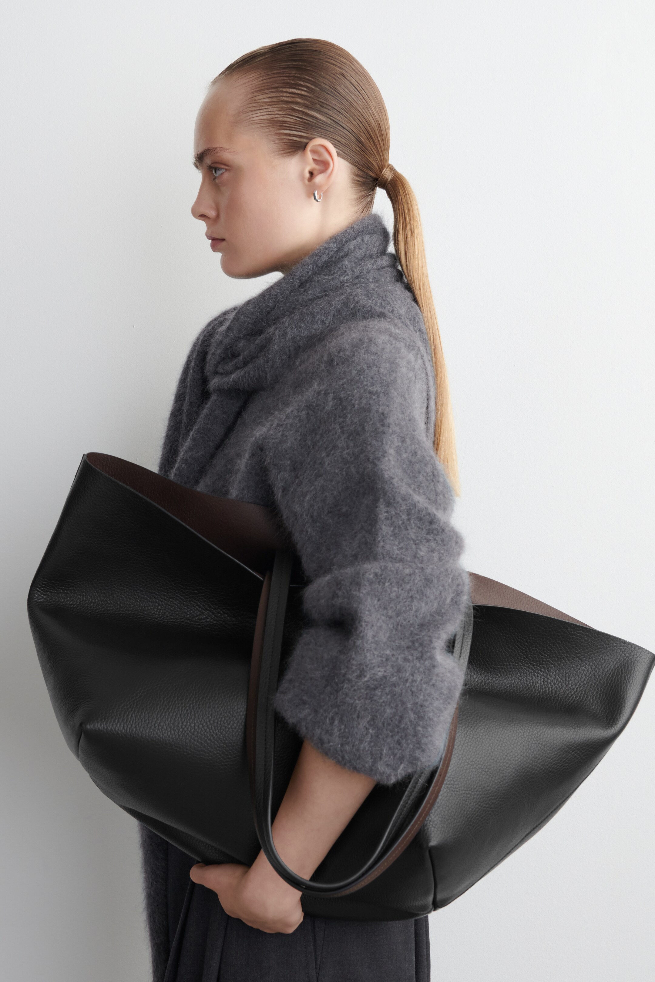 Agrandir l'image: Un grand sac fourre-tout noir, fabriqué en cuir à grain doux, est tenu du côté GAUCHE. Sa forme ouverte révèle une doublure marron foncé contrastante, et des fines sangles sombres s'étendent vers le haut.