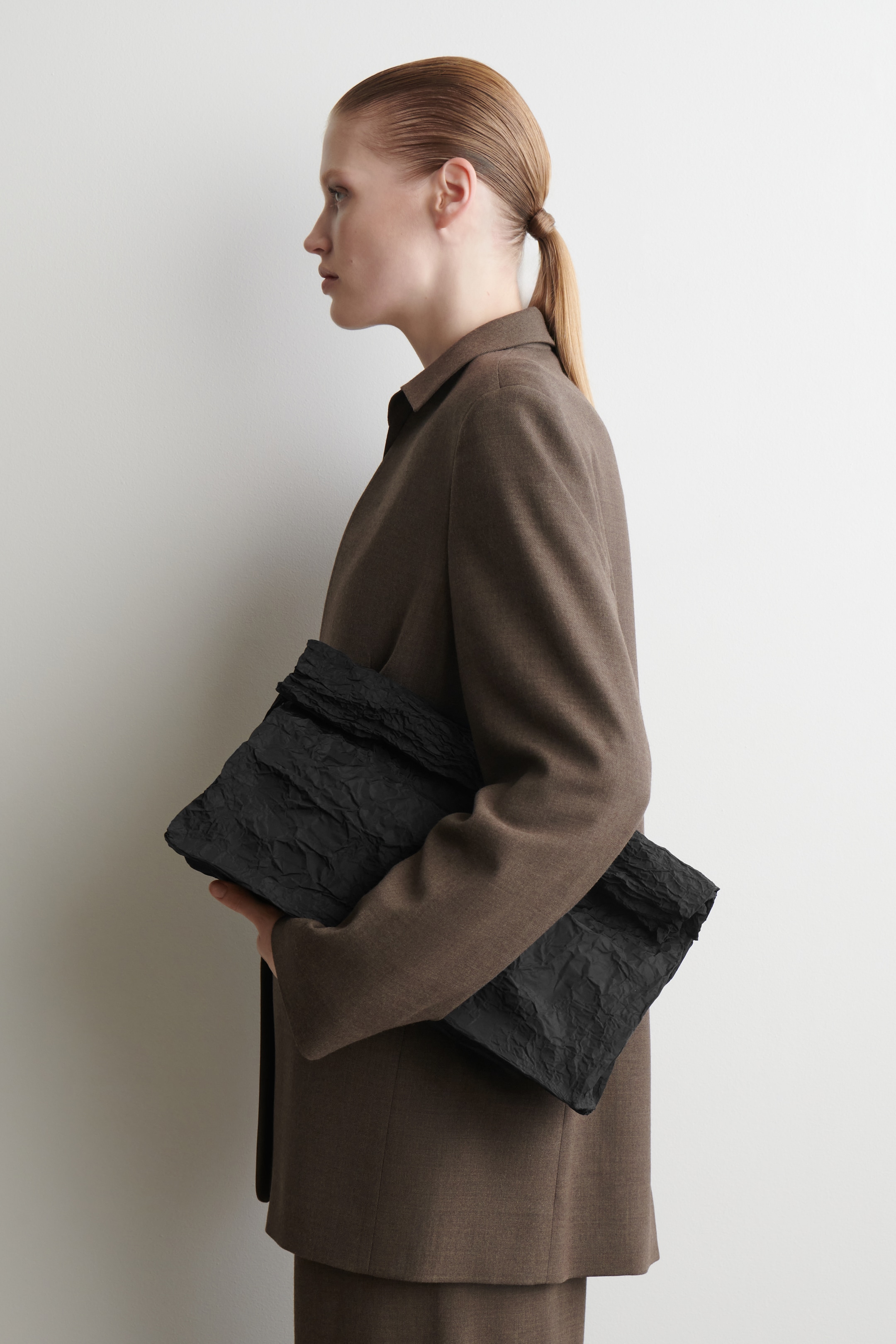 Visualizza immagine più grande: Una borsa a mano nera oversize viene tenuta orizzontalmente sotto il braccio sinistro di una persona, rivolta a sinistra, caratterizzata da una texture stropicciata pronunciata e un top esagerato e ripiegato.