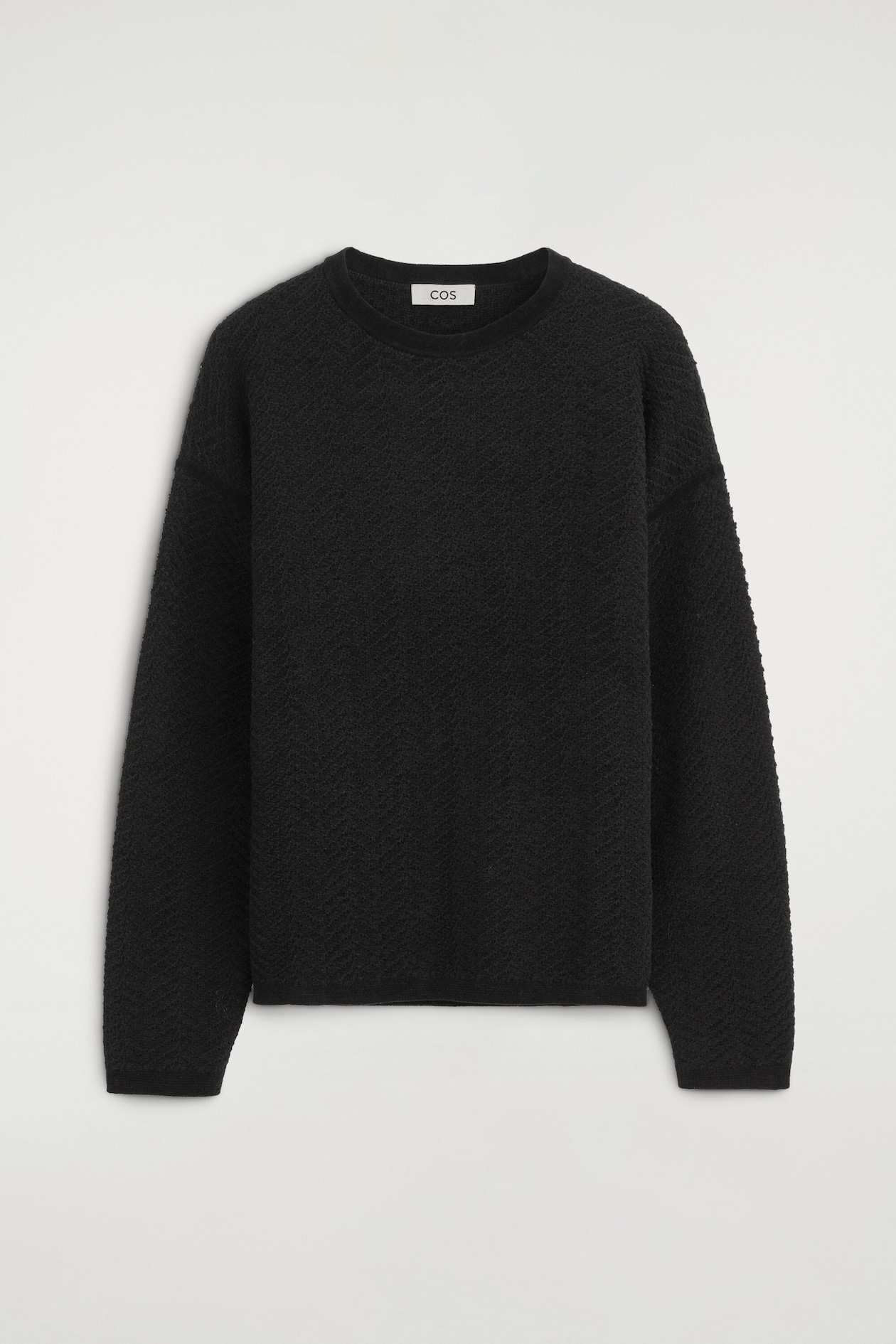 The merino wool chenille crew-neck sweater is shown from the front, highlighting its textured black knit with a subtle diagonal pattern and dropped shoulders. It features a ribbed crew neckline, long sleeves with ribbed cuffs, and a ribbed hem.