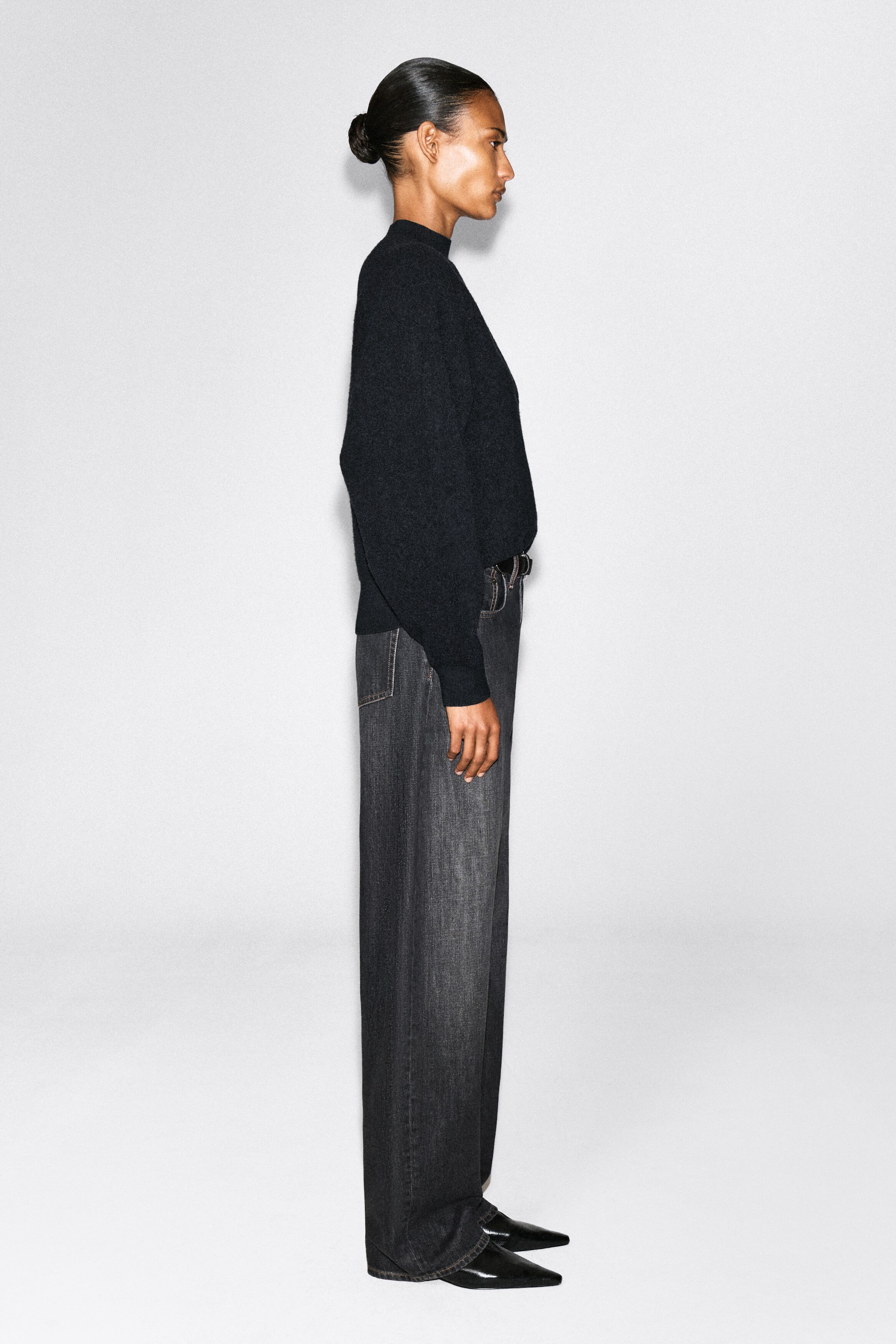 The model is shown in profile wearing a black mock-neck knit sweater and dark gray wide-leg jeans. The long-sleeve sweater is tucked into the waist, and the high-waisted jeans drape over black pointed-toe shoes. The model is shown in profile wearing a black mock-neck knit sweater and dark gray wide-leg jeans. The long-sleeve sweater is tucked into the waist, and the high-waisted jeans drape over black pointed-toe shoes.