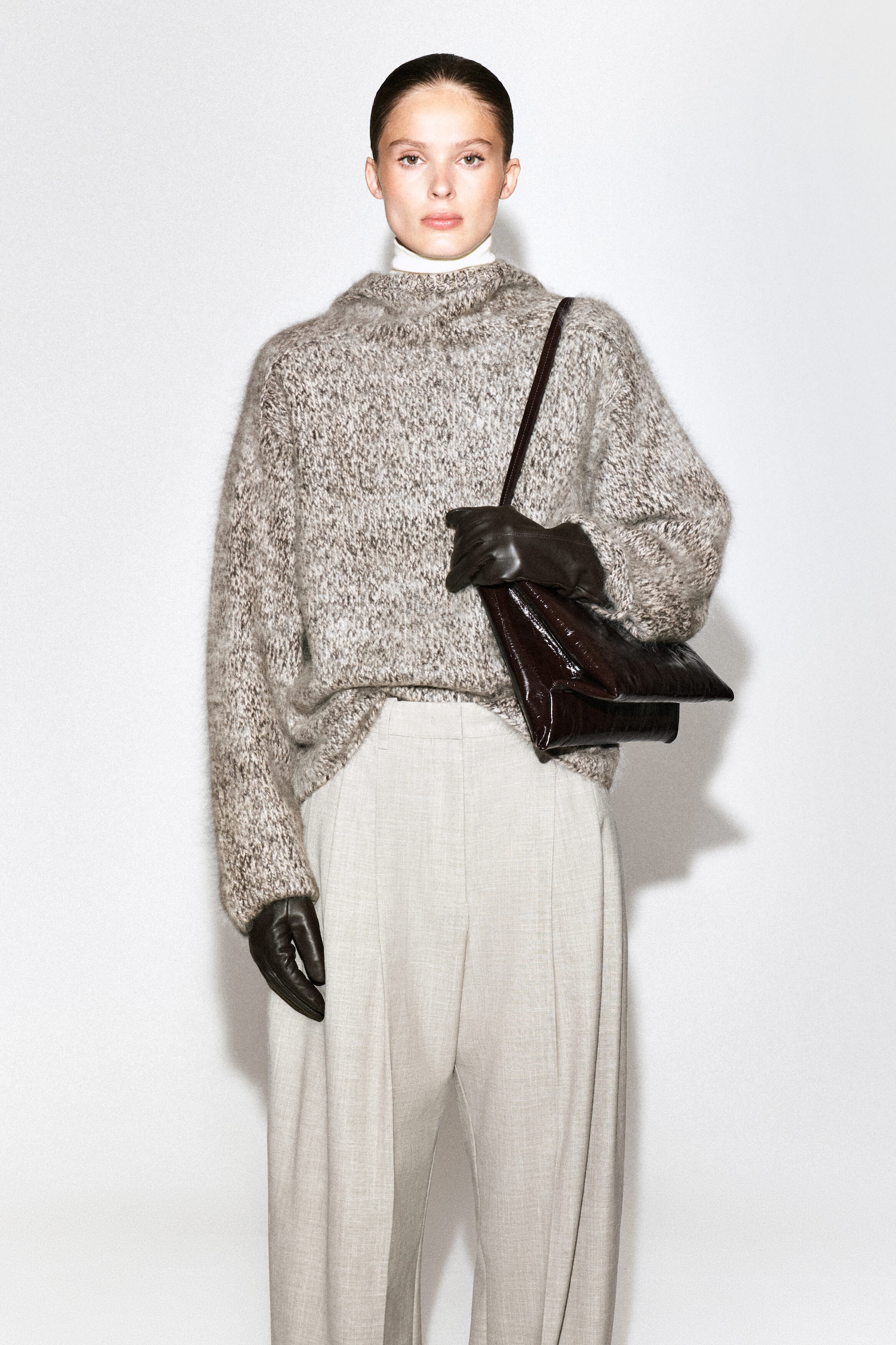 Un chandail en tricot chiné gris et brun à motif moucheté, avec un col montant et des manches longues, est porté par le mannequin. Le chandail surdimensionné est agencé avec un pantalon large plissé de couleur pâle, tandis que des gants en cuir brun foncé sont portés et qu’un sac à épaule assorti en cuir brun foncé est transporté. Un chandail en tricot chiné gris et brun à motif moucheté, avec un col montant et des manches longues, est porté par le mannequin. Le chandail surdimensionné est agencé avec un pantalon large plissé de couleur pâle, tandis que des gants en cuir brun foncé sont portés et qu’un sac à épaule assorti en cuir brun foncé est transporté.